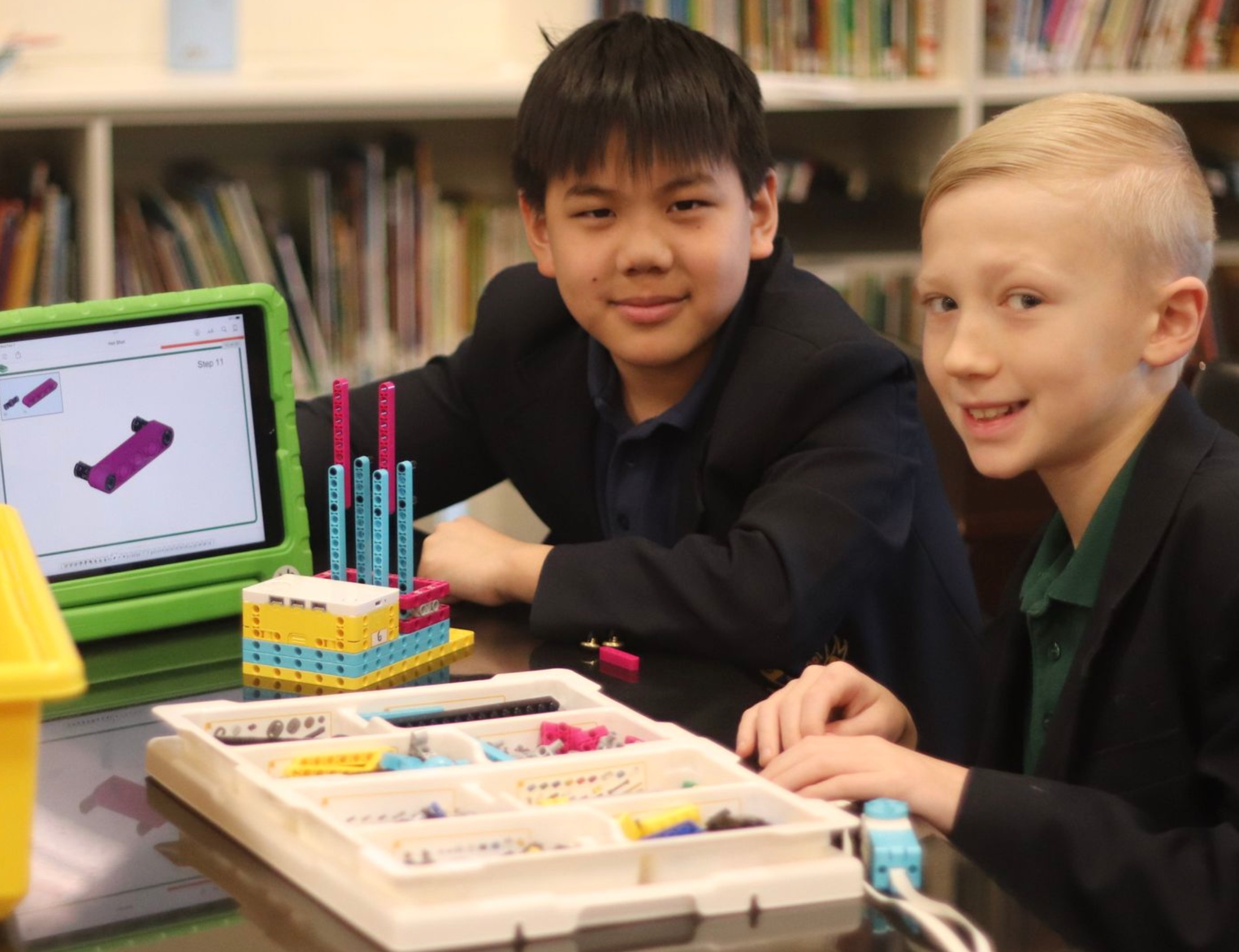 Two boys sitting at a table with robotics building kits and a tablet, smiling at the camera, in a library or classroom setting.