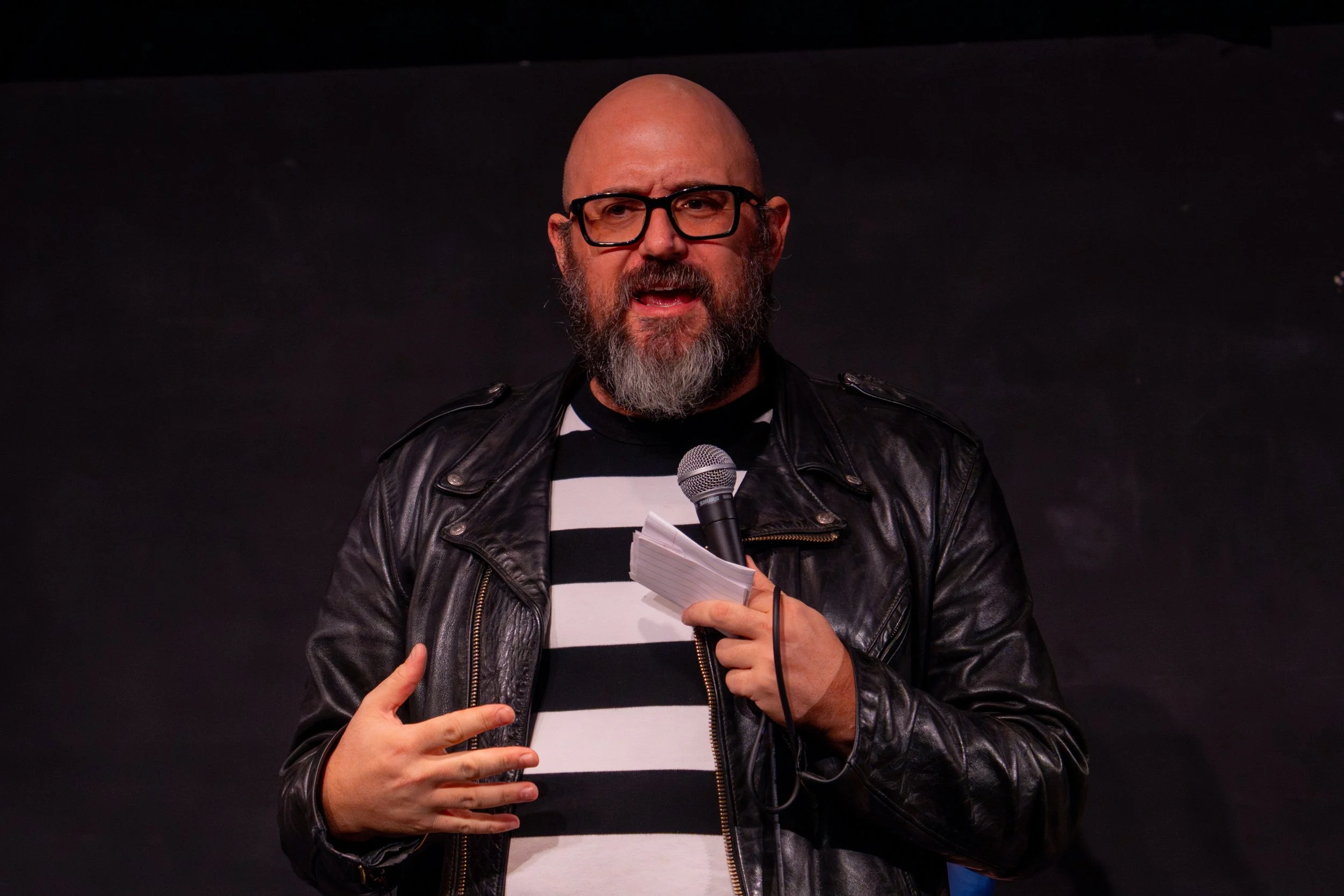 Jeff Simmermon performs CareGiver at SoHo Playhouse, speaking into a handheld microphone and wearing a black leather jacket over a striped shirt.