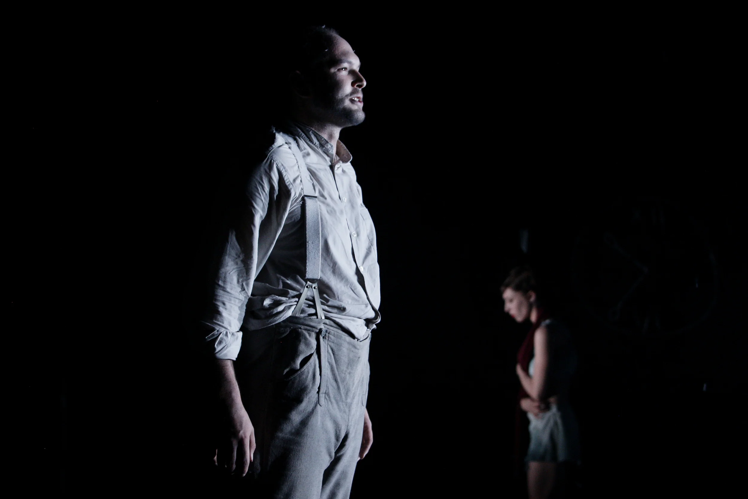 A man and a woman standing in a dark room with dramatic lighting, the man in the foreground gazing upward, the woman in the background looking down. From Heather Fairbairn's operatic adaptation of Schubert's Winterreise.