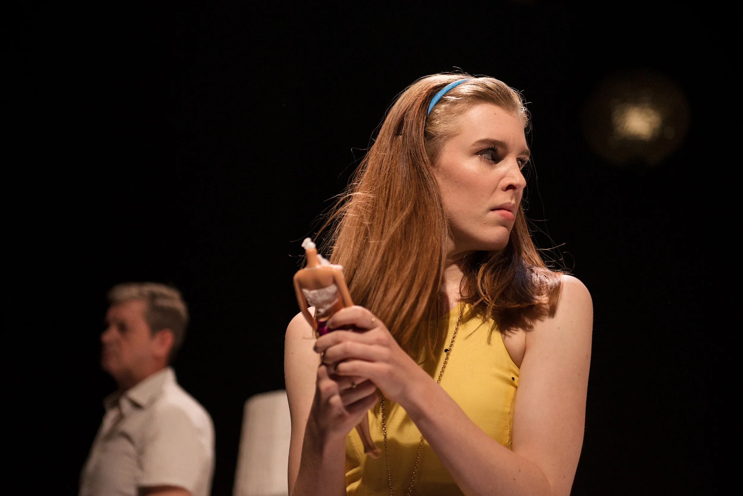 A woman with red hair and a blue headband sitting on stage, holding a Barbie doll, with a man blurred in the background. From Sunnytown, directed by Heather Fairbairn.