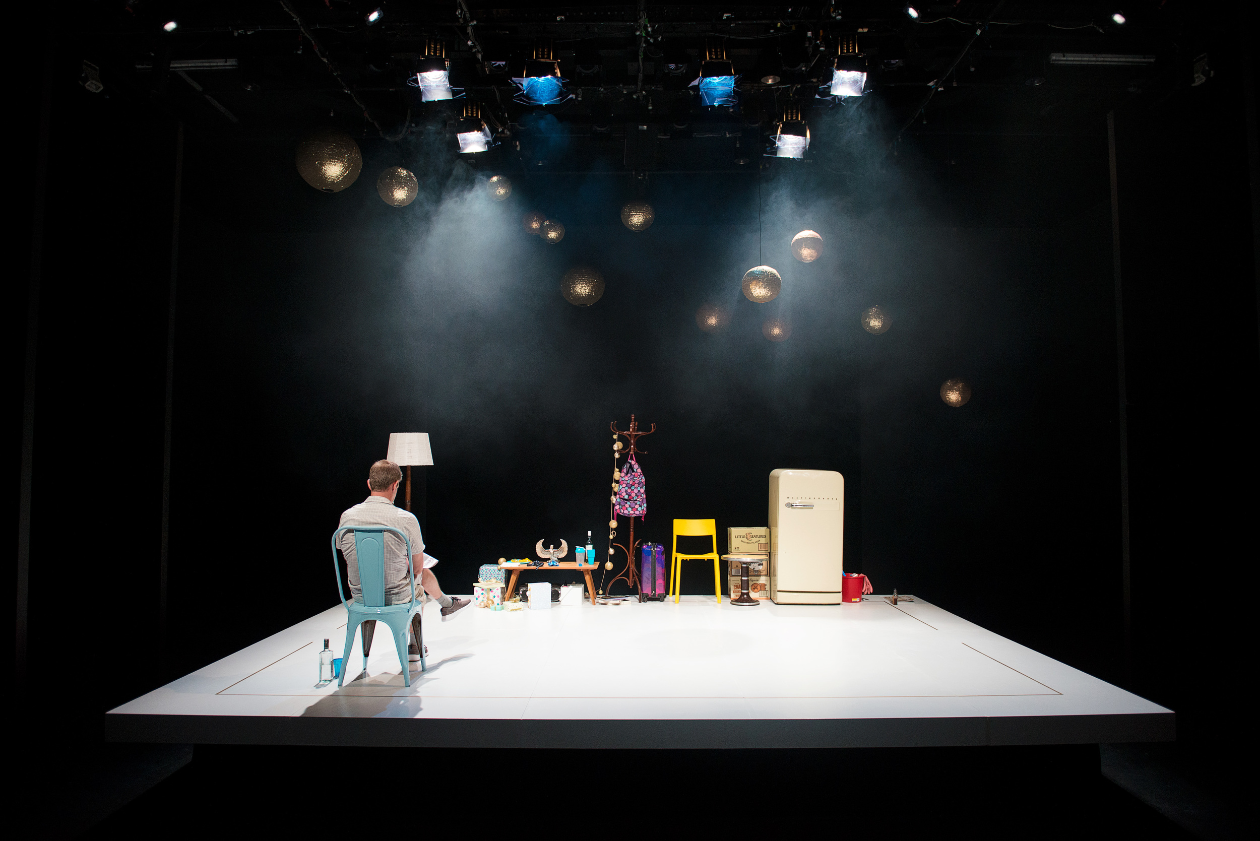 A theater stage set with a man sitting on a chair facing away, surrounded by vintage furniture and household items, with hanging spherical lights and stage lighting overhead. From Sunnytown, directed by Heather Fairbairn.