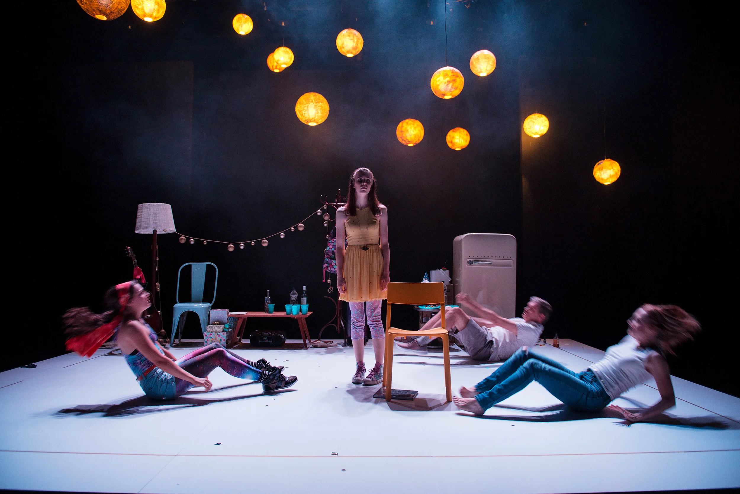 Stage performance with four young women; one standing in yellow dress, three sitting or laying on floor; set includes hanging orange lanterns, table with drinks, lamp, chairs, refrigerator, and string lights.