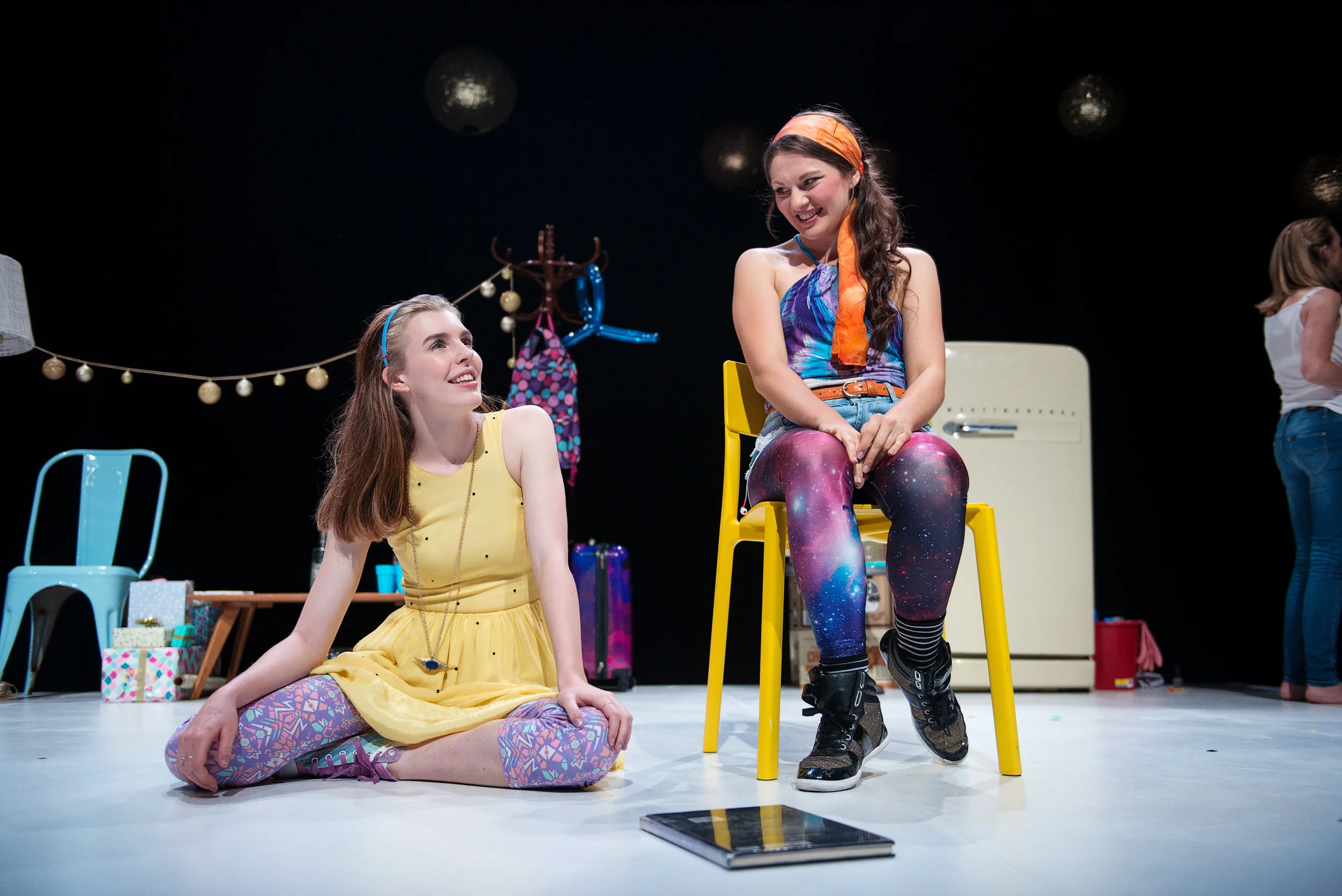 Two young women on stage, one sitting cross-legged on the floor in a yellow dress and the other sitting on a yellow chair in galaxy-patterned leggings and a colorful top, engaging in conversation with smiles and relaxed posture. Stage props include a