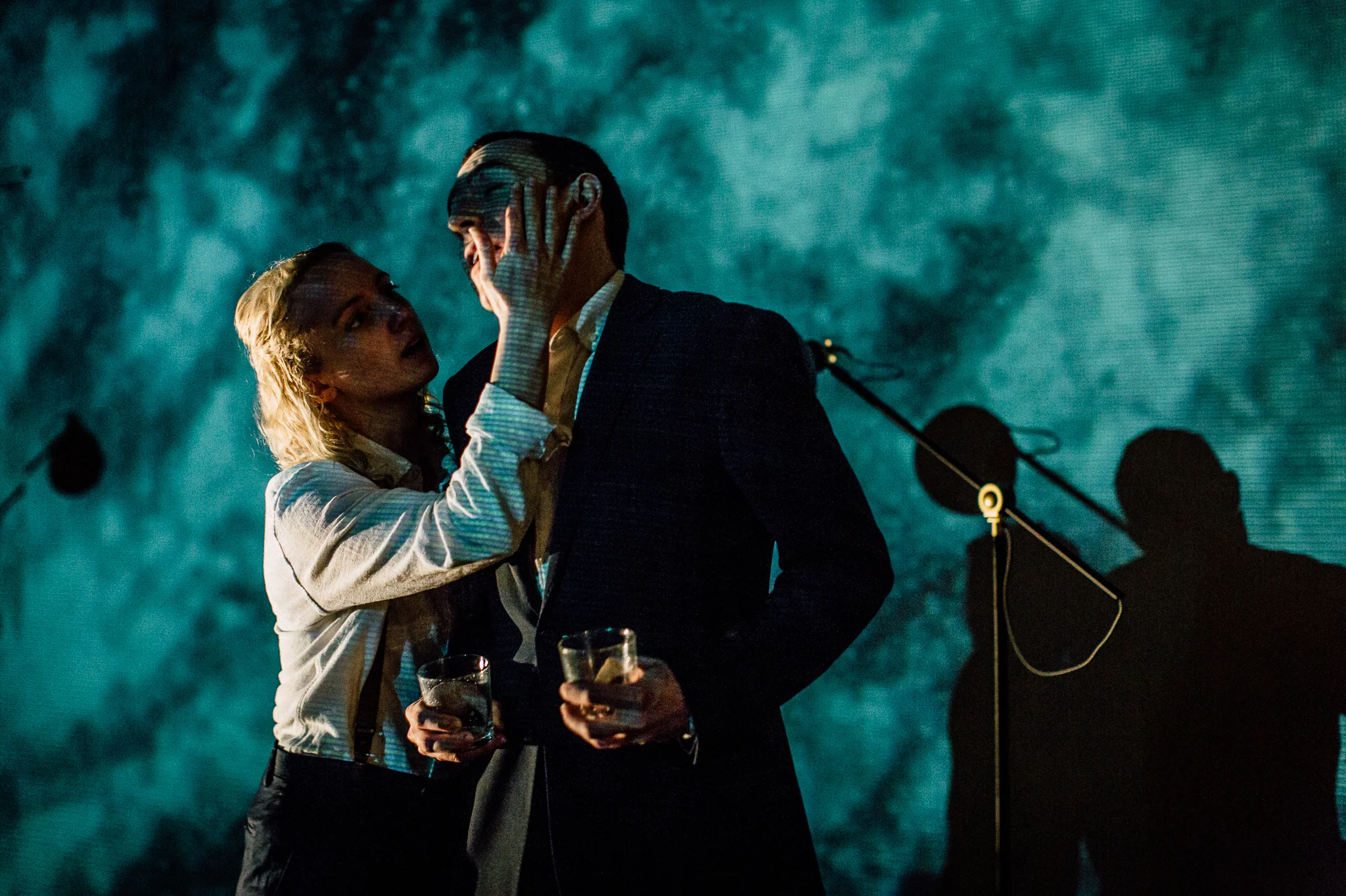 A man and a woman look at each other closely, with the woman's hand on the man's face. They are holding glasses and are dressed formally, standing on a stage with microphone stands and speakers in the background under moody lighting.