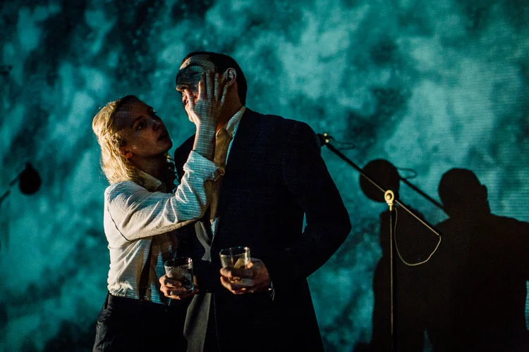 A woman touches the face of a man in a suit holding a glass, on a stage with microphone stand and shadows in the background. From the play The Mutant Man, directed by Heather Fairbairn at The Space Arts Centre, London.