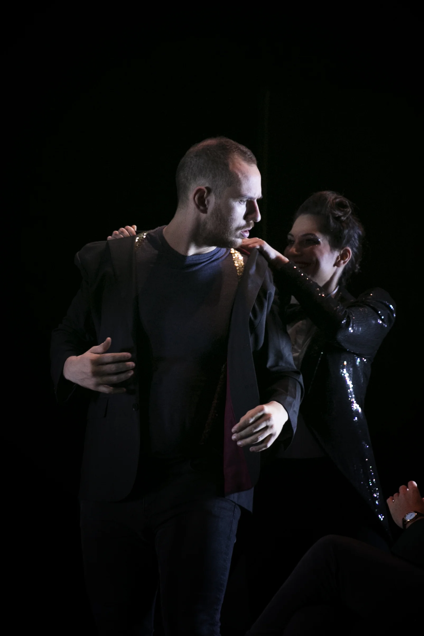 Heather Fairbairn's production of Ariadne auf Naxos. A man and woman performing on stage, with the woman touching the man's shoulder and smiling, both dressed in dark clothing with shiny accents, against a black background.
