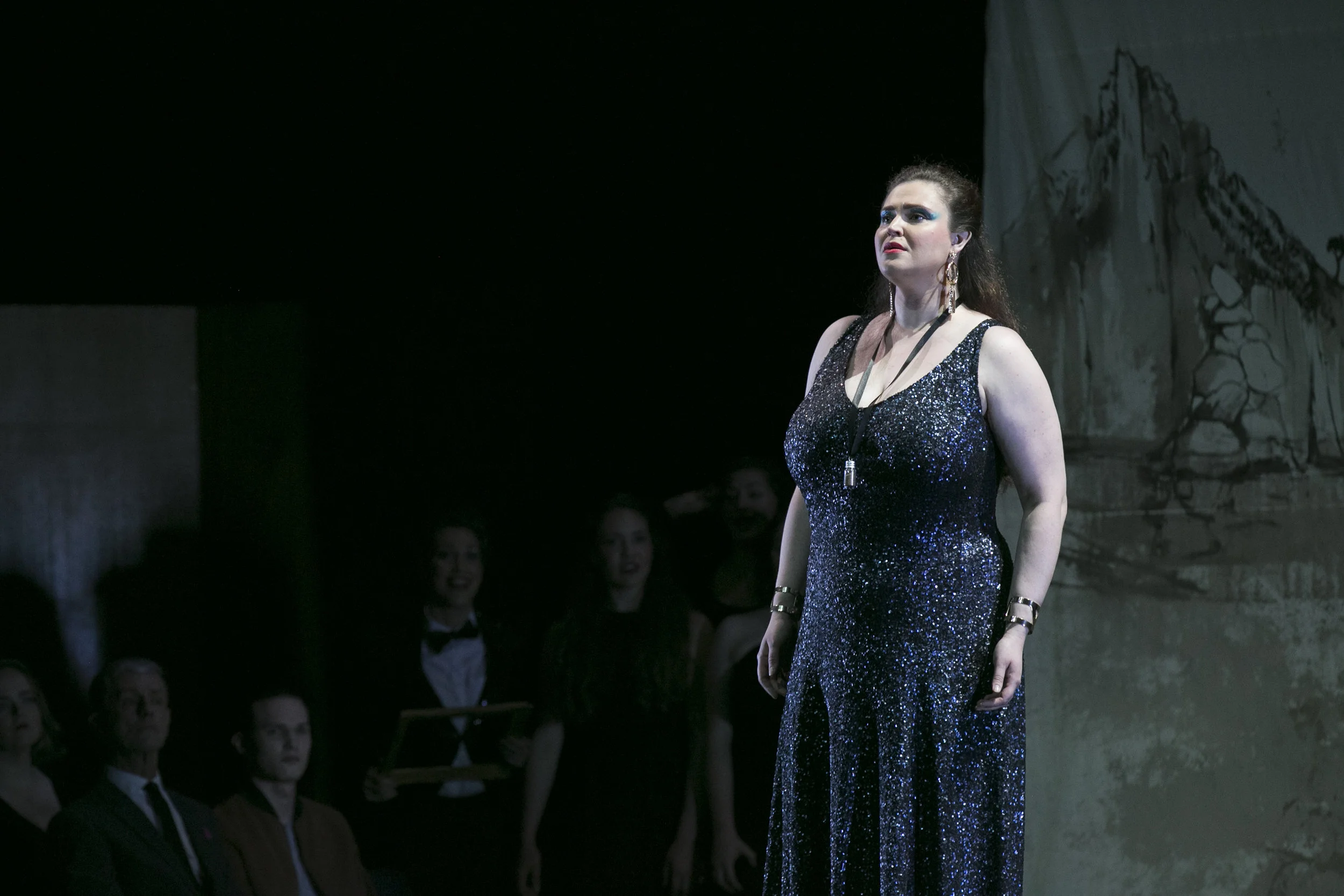 A woman wearing a sparkly black dress stands on stage with a serious expression, facing forward. Several people, some in formal attire, are in the background watching her, with a painted backdrop featuring mountains behind her.