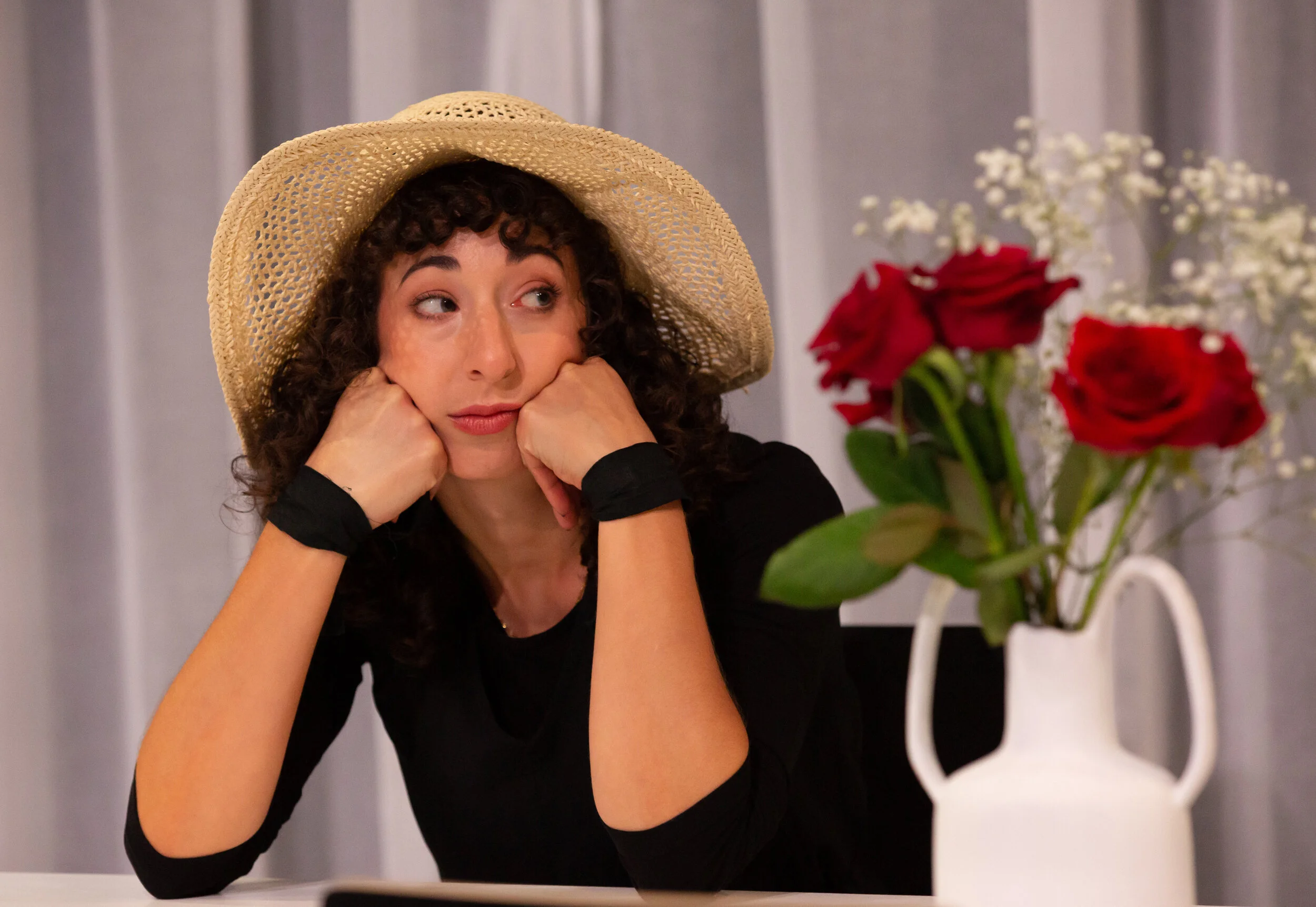 Woman with curly hair wearing a straw hat, resting her chin on her hands, sitting at a table with a vase of red roses and white flowers, and looking to the side with a bored or annoyed expression. From director Heather Fairbairn's Elektra/Orestes.