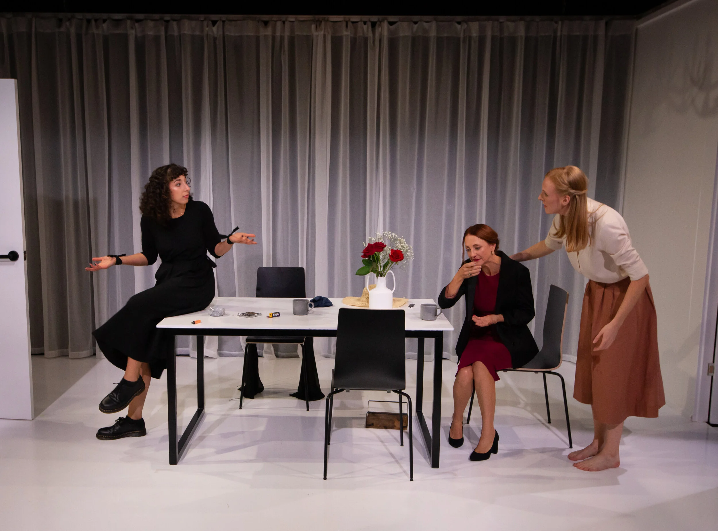 Three women engaged in a conversation around a white table with a vase of red roses, set with cups, snacks, and personal items. One woman with curly dark hair is sitting on the table, gesturing with her hands. Another woman with red hair, wearing a b
