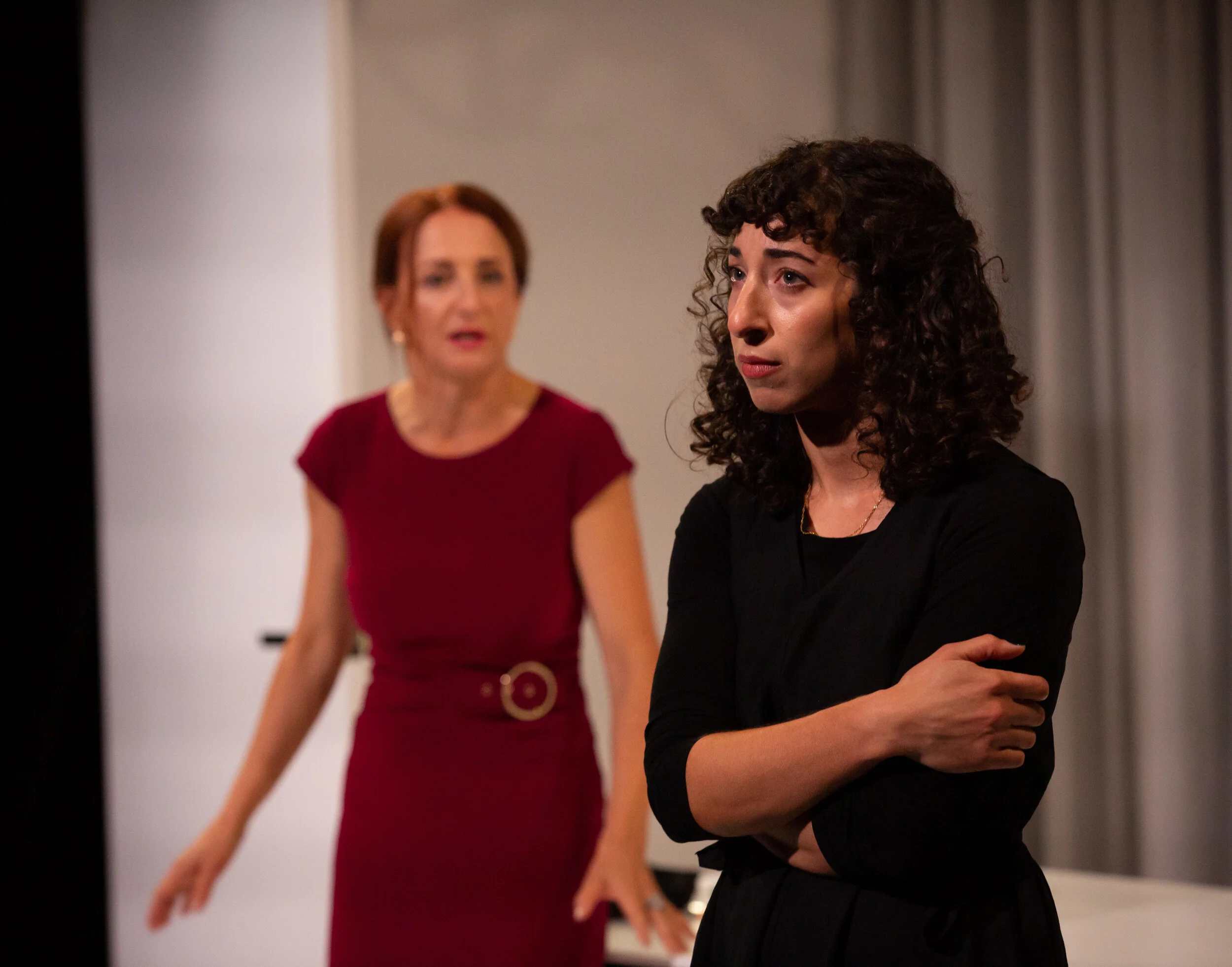 Two women in conversation, one in the foreground with arms crossed and a serious expression, the other in the background gesturing and talking, both indoors with curtains behind them. From director Heather Fairbairn's production of Elektra/Orestes.