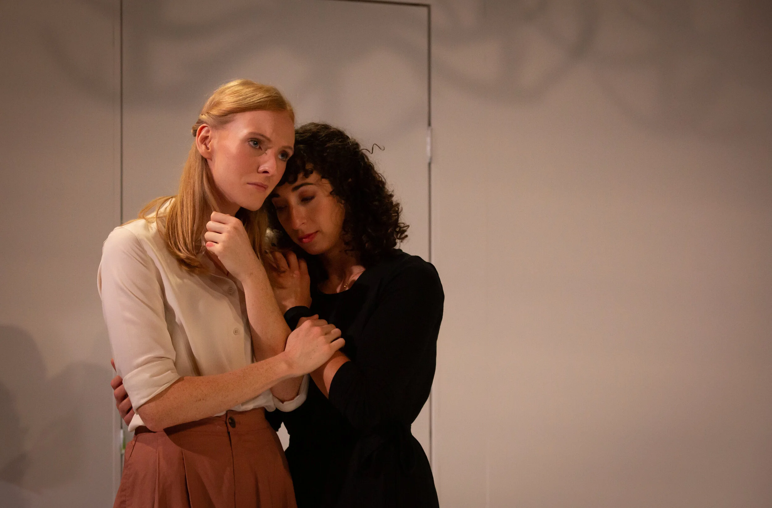 Two women standing closely together, showing concern or comforting each other in front of a plain wall. From director Heather Fairbairn's production of Elektra/Orestes.