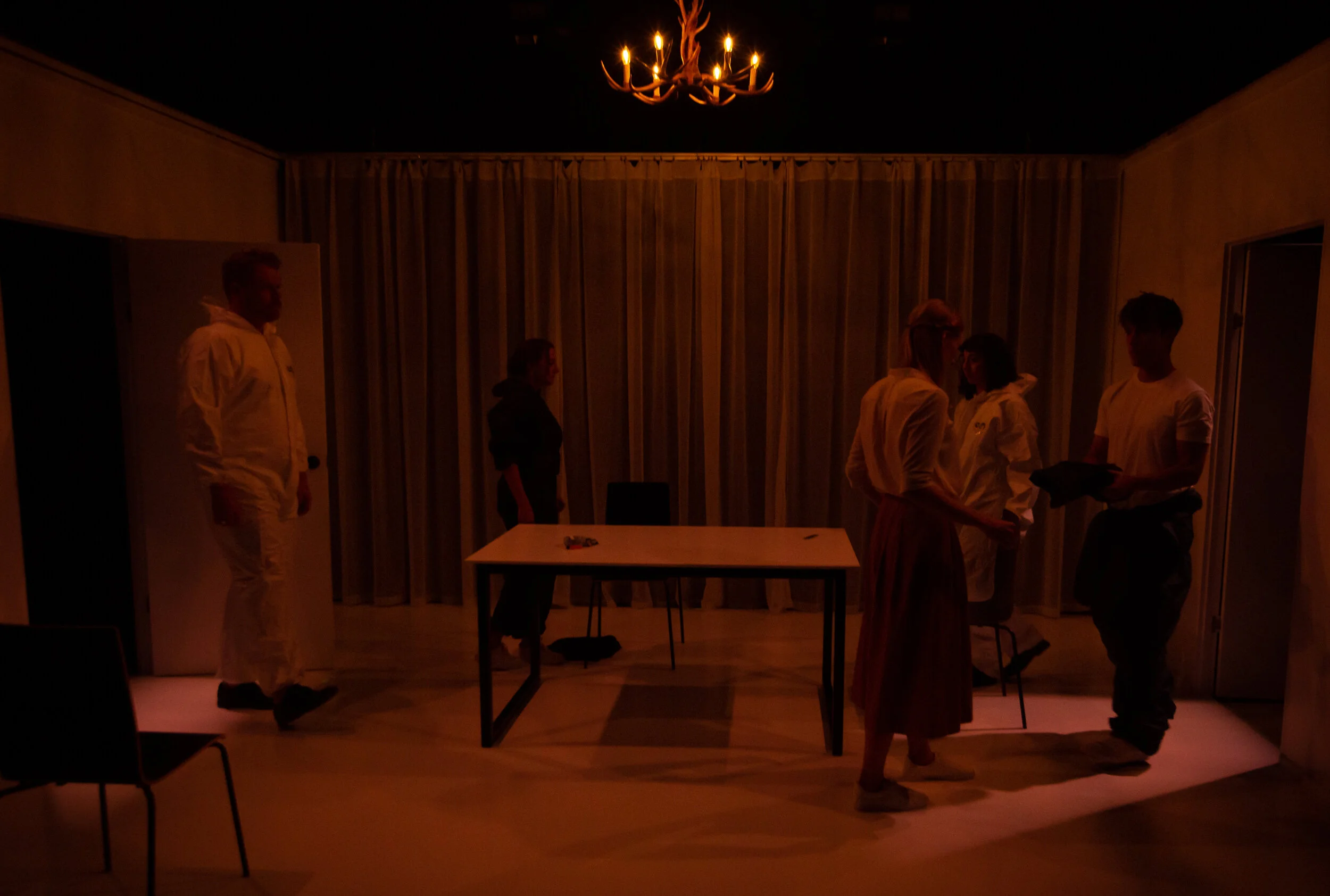 Six people in a dimly lit room with a chandelier, some near a table, engaged in conversation or activity. From director Heather Fairbairn's production of Elektra/Orestes.