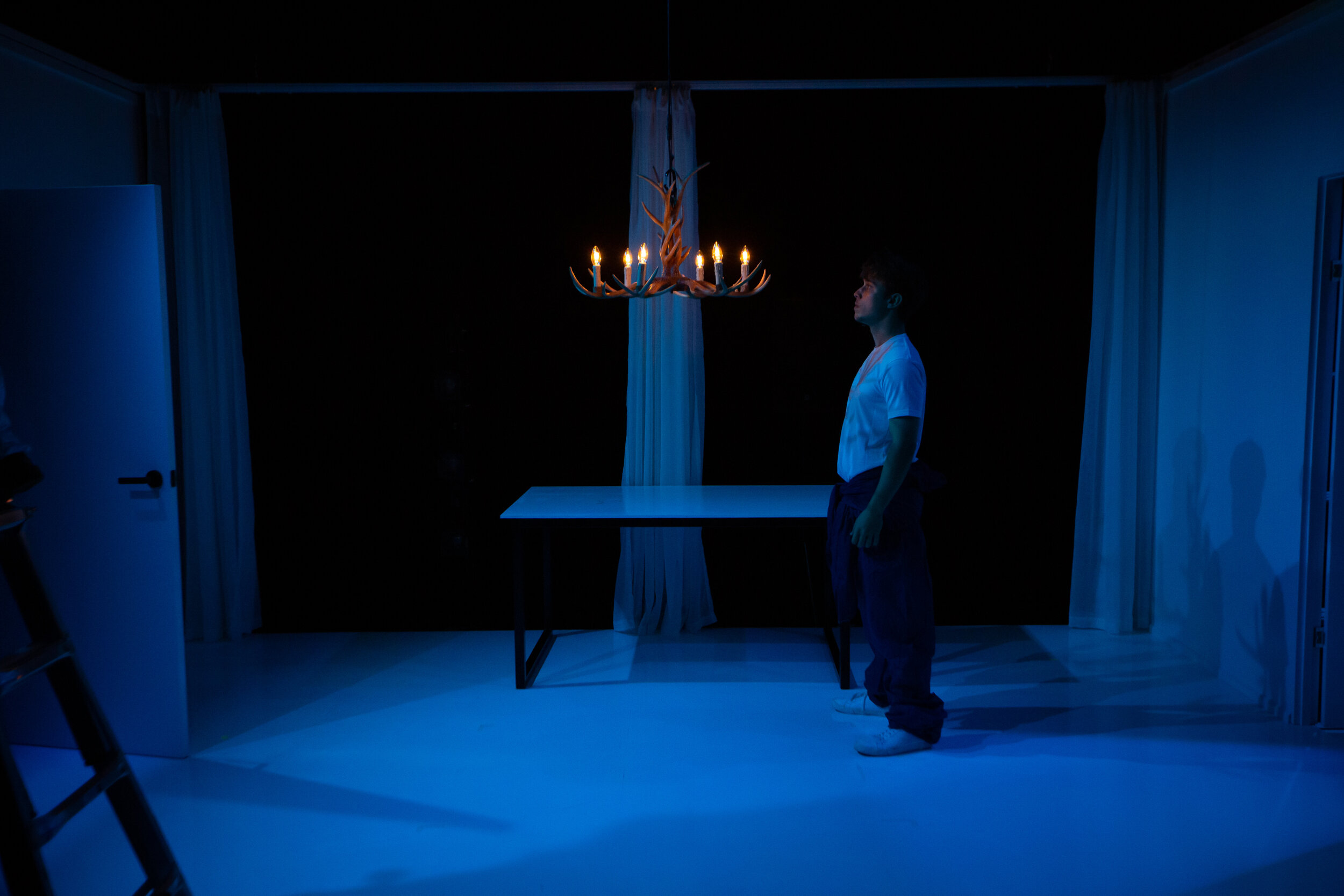 A young man in a white shirt and dark pants standing in a dimly lit room, facing a table with a curtain behind it, illuminated by a chandelier with lit candles hanging from the ceiling. From director Heather Fairbairn's production of Elektra/Orestes.
