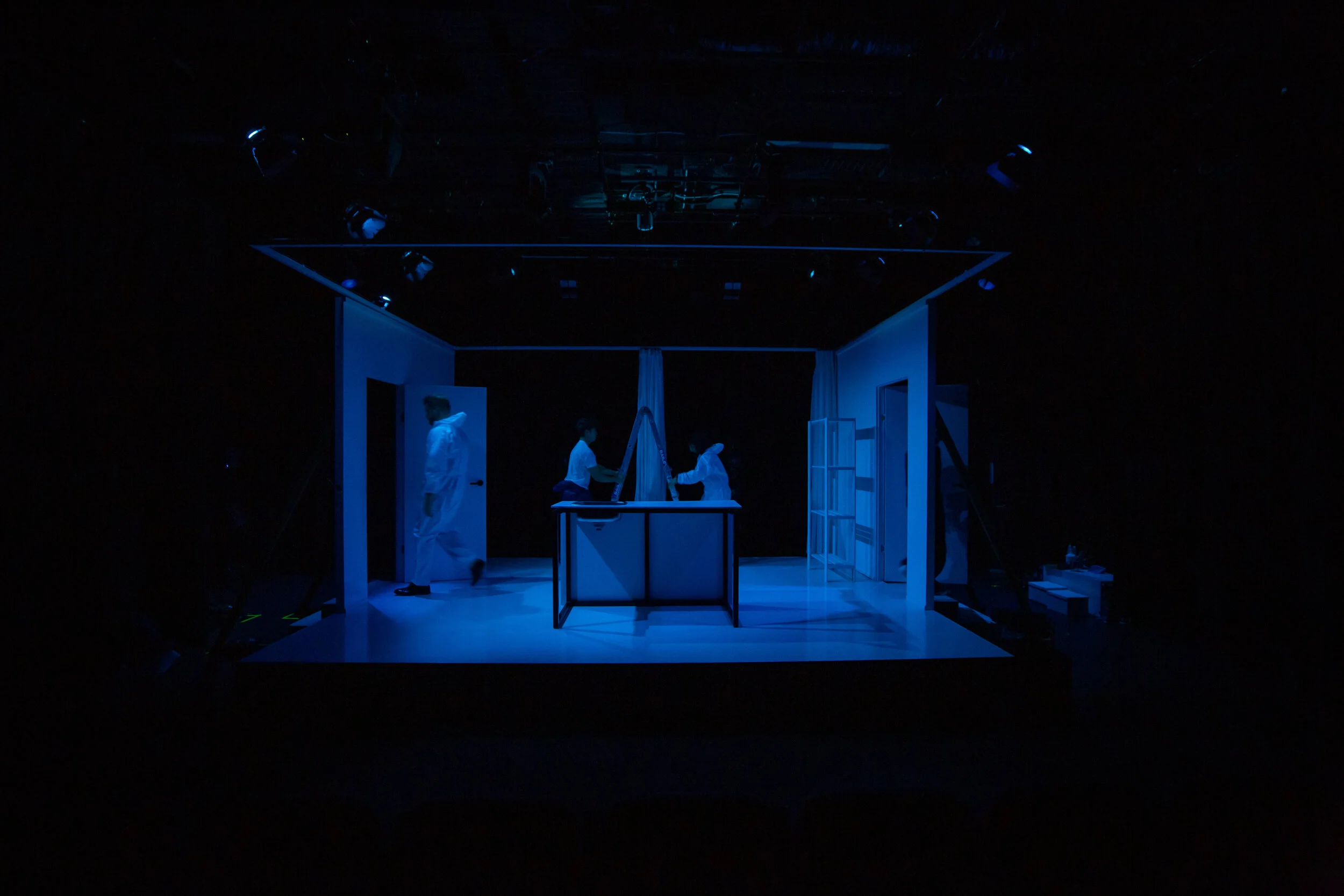 A stage with a small set designed to resemble a room, illuminated by blue lighting. Four people dressed in white are on stage, some holding the curtain and others walking or standing around. From director Heather Fairbairn's Elektra/Orestes.
