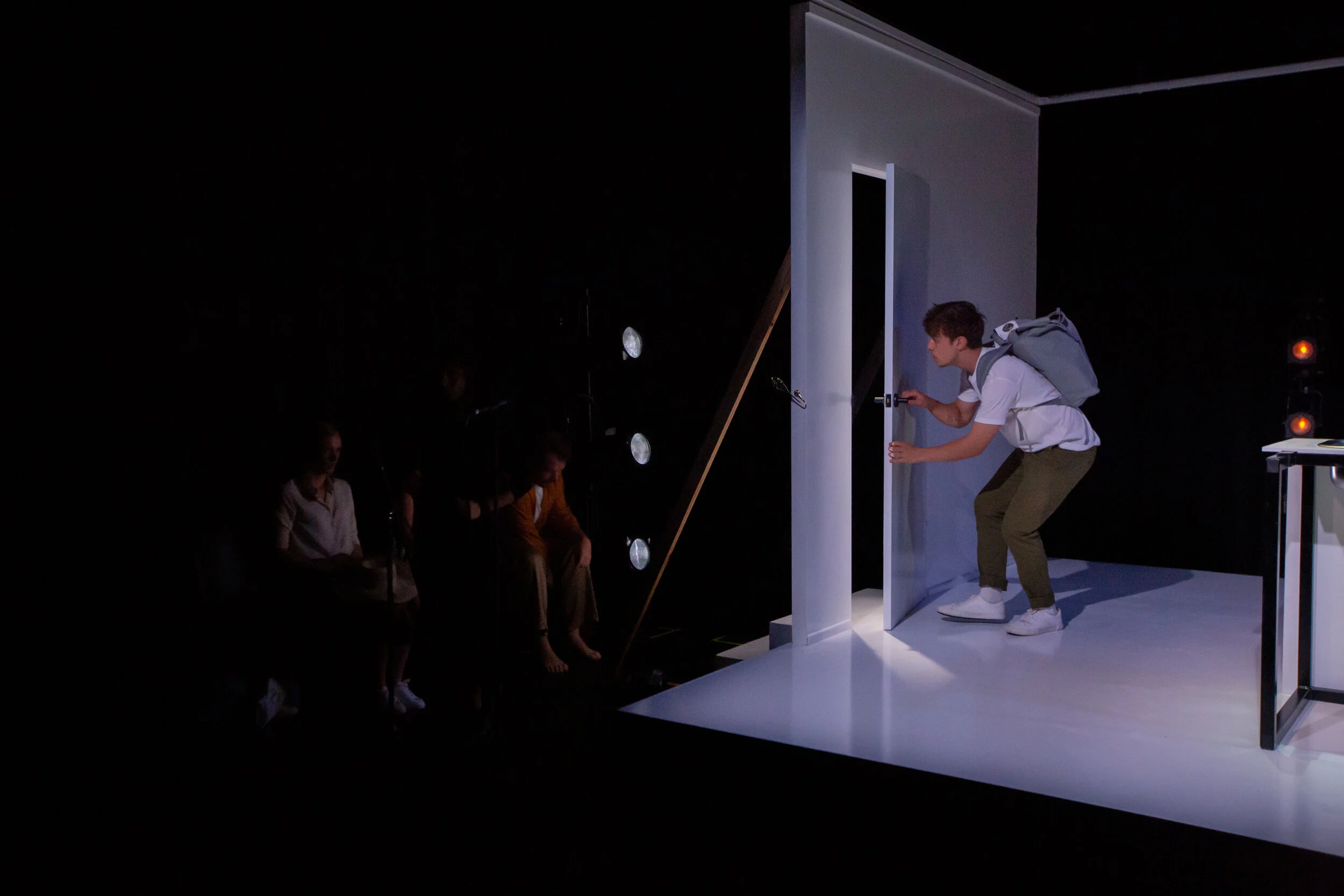 A young man with a backpack peeking through a door onto an audience in a dark theater stage. From director Heather Fairbairn's production of Elektra/Orestes.