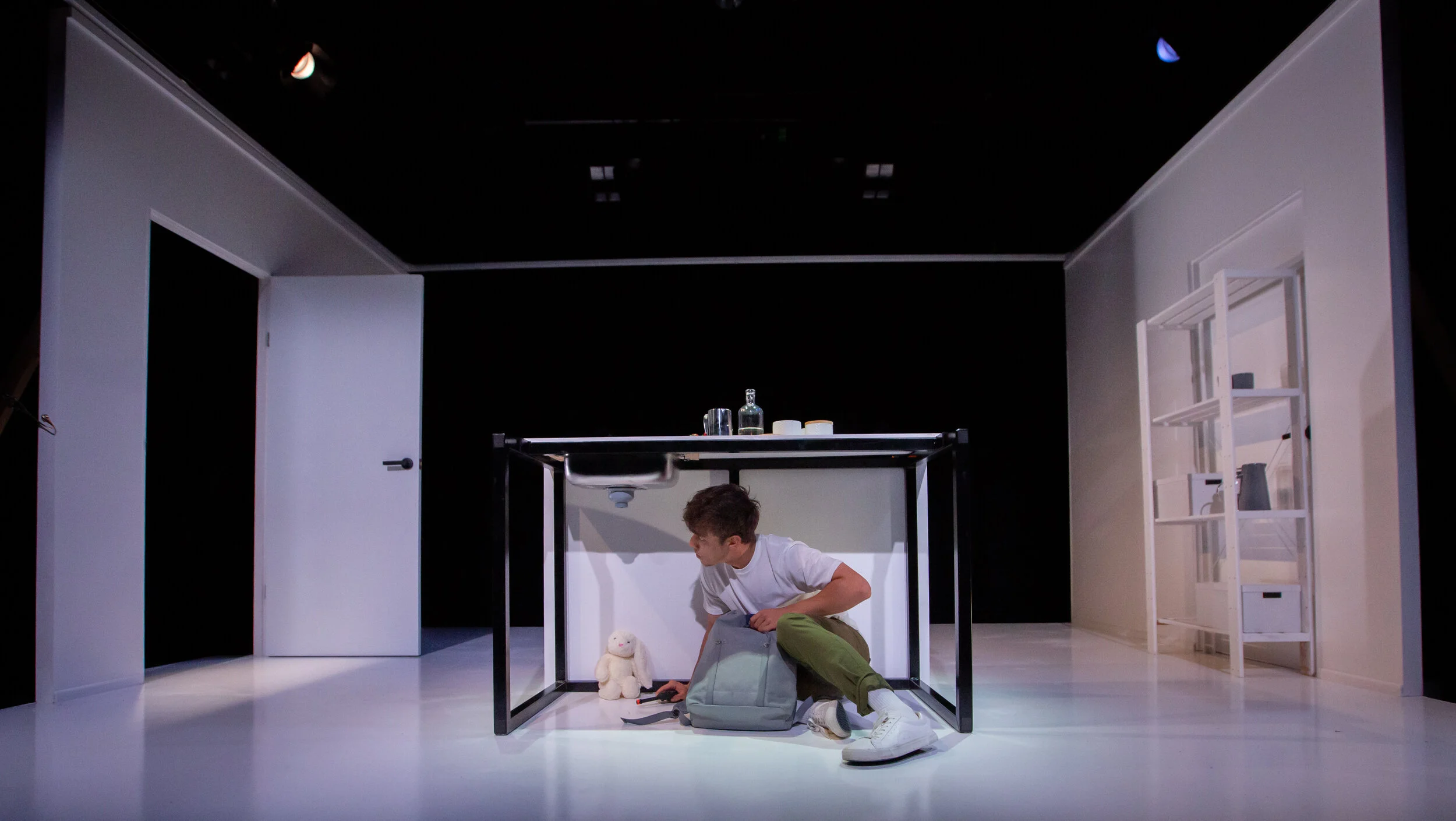 A young man in a white T-shirt and green pants sitting under a black kitchen table in a white room, reaching into a gray backpack, with a white plush toy and a black device on the floor. The room has a white door on the left and a white shelving unit