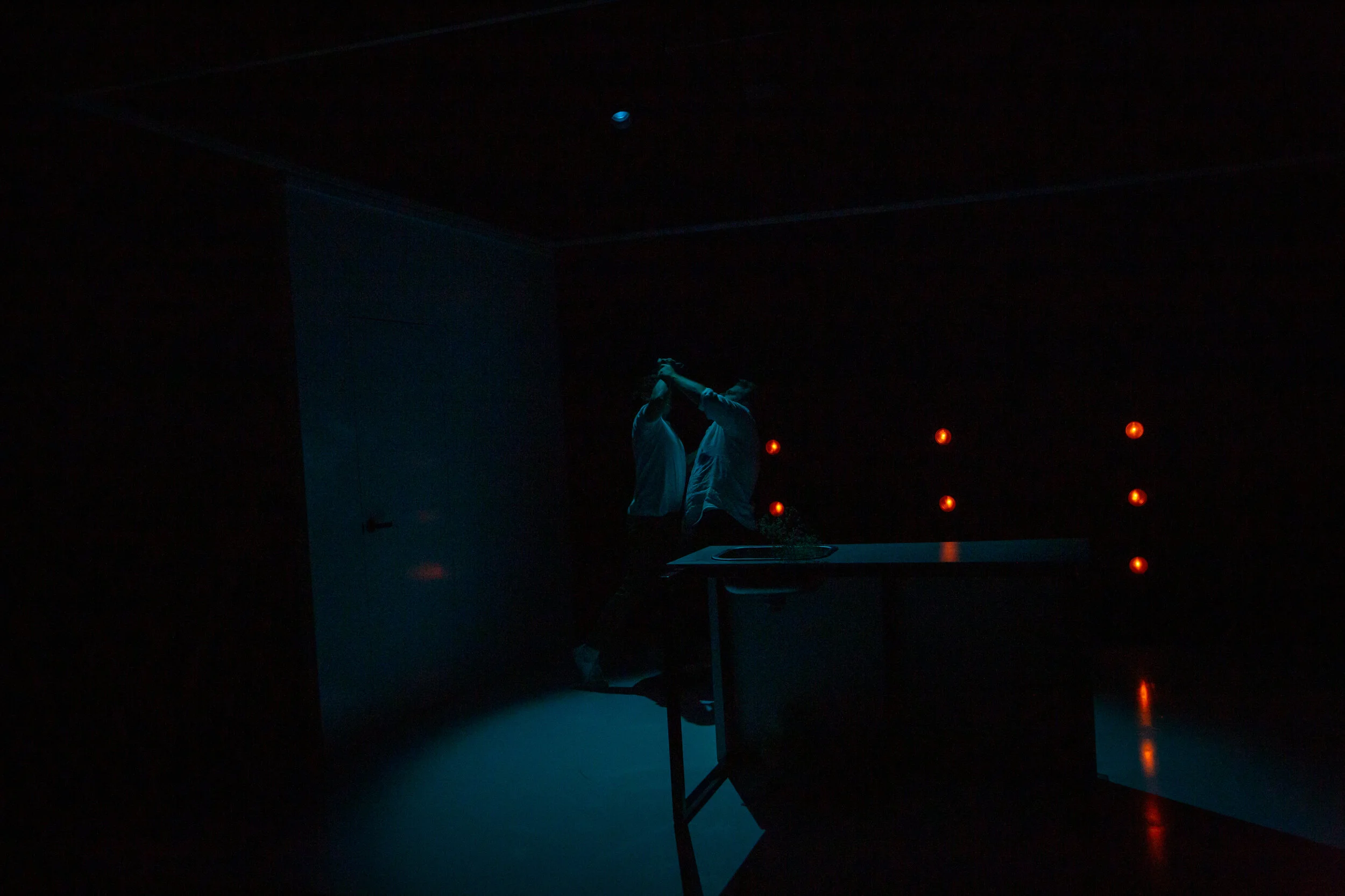 Heather Fairbairn's production of Elektra/Orestes. Two people in white shirts dancing in a dark room with red lights on the wall, illuminated slightly by low lighting.