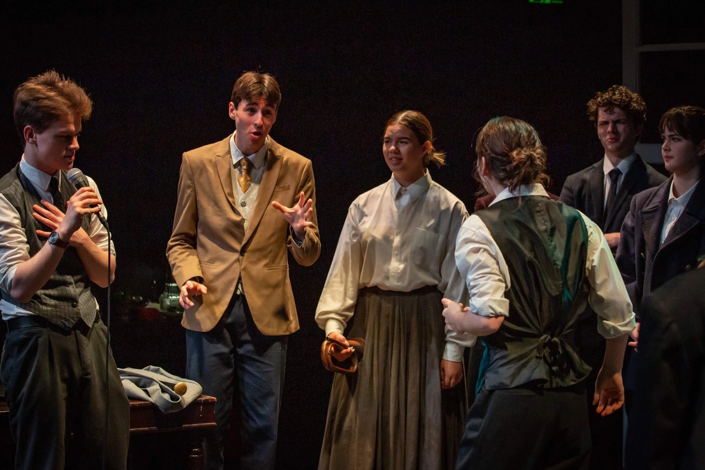 Heather Fairbairn's production of An Enemy of the People at Queensland Theatre. Group of young people performing on stage during a theatrical play, with one person speaking into a microphone and others listening.