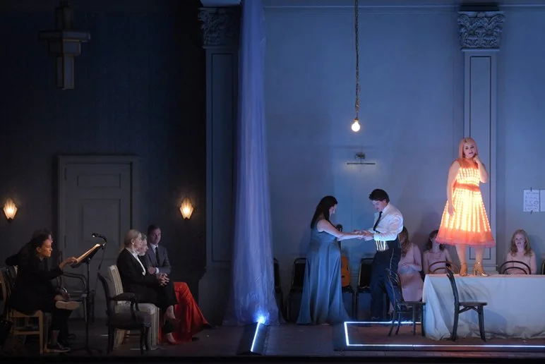 A theatrical performance on stage with actors and a puppet. One woman stands on a table dressed in a light pink dress with glowing effect, while a man and a woman are exchanging a document or paper in the center. Several people sit watching, some wit