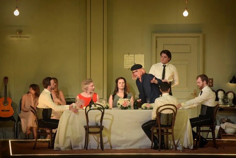 People seated around a dining table in a room with green walls, dim lighting, and guitars in the background, with someone being held by another person near the center.