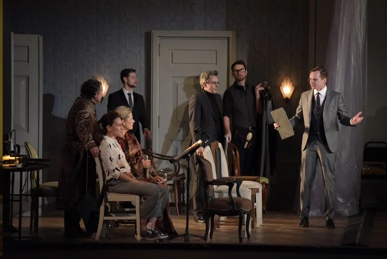 Scene from a theatrical play with seven actors on stage, some seated and some standing, in a room with a door and vintage furniture, with a director or speaker gesturing.