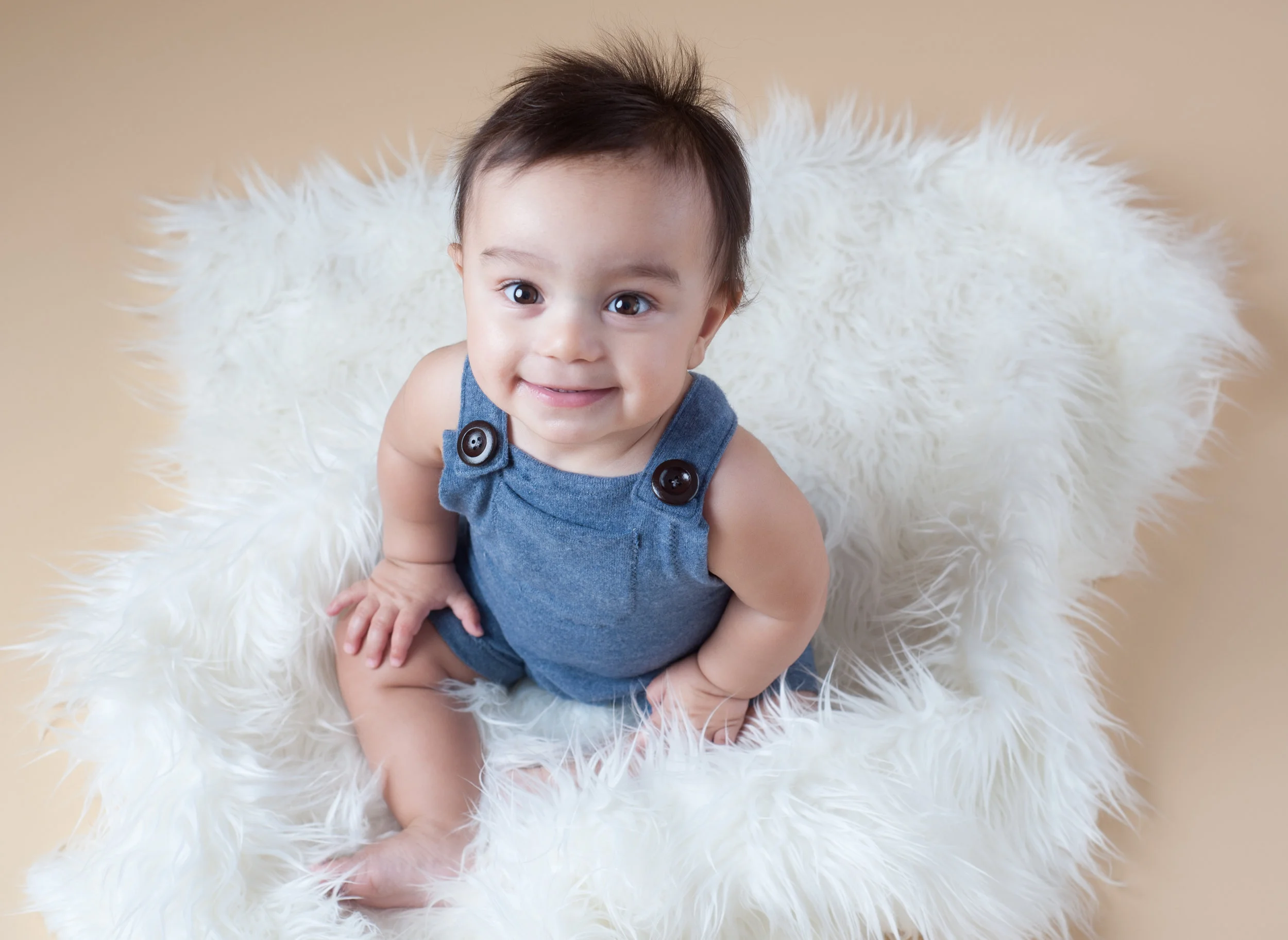 Baby boy in a blue romper sitting up on a white rug and looking up smiling for his sitter session