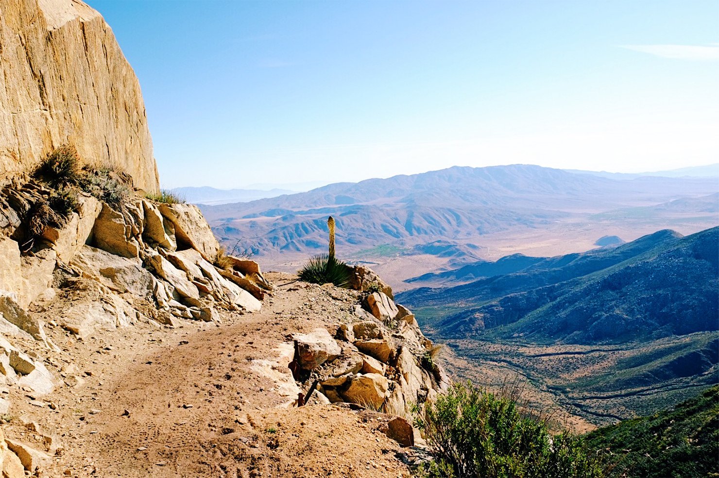 A dirt trail along a rocky mountainside with a desert landscape and mountains in the distance, under a clear blue sky.