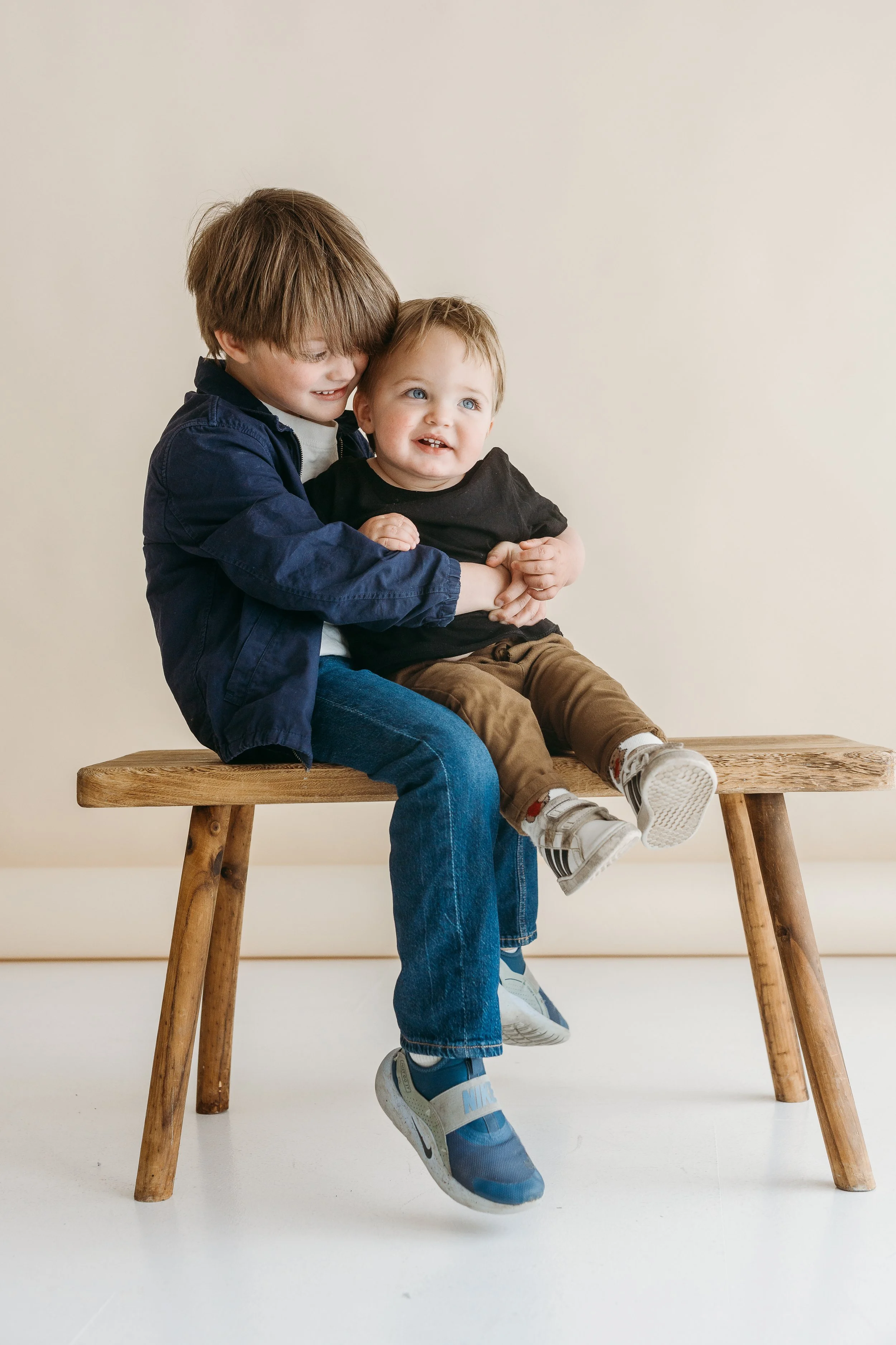 Two young boys sitting on a wooden bench, smiling and hugging each other against a plain beige wall.