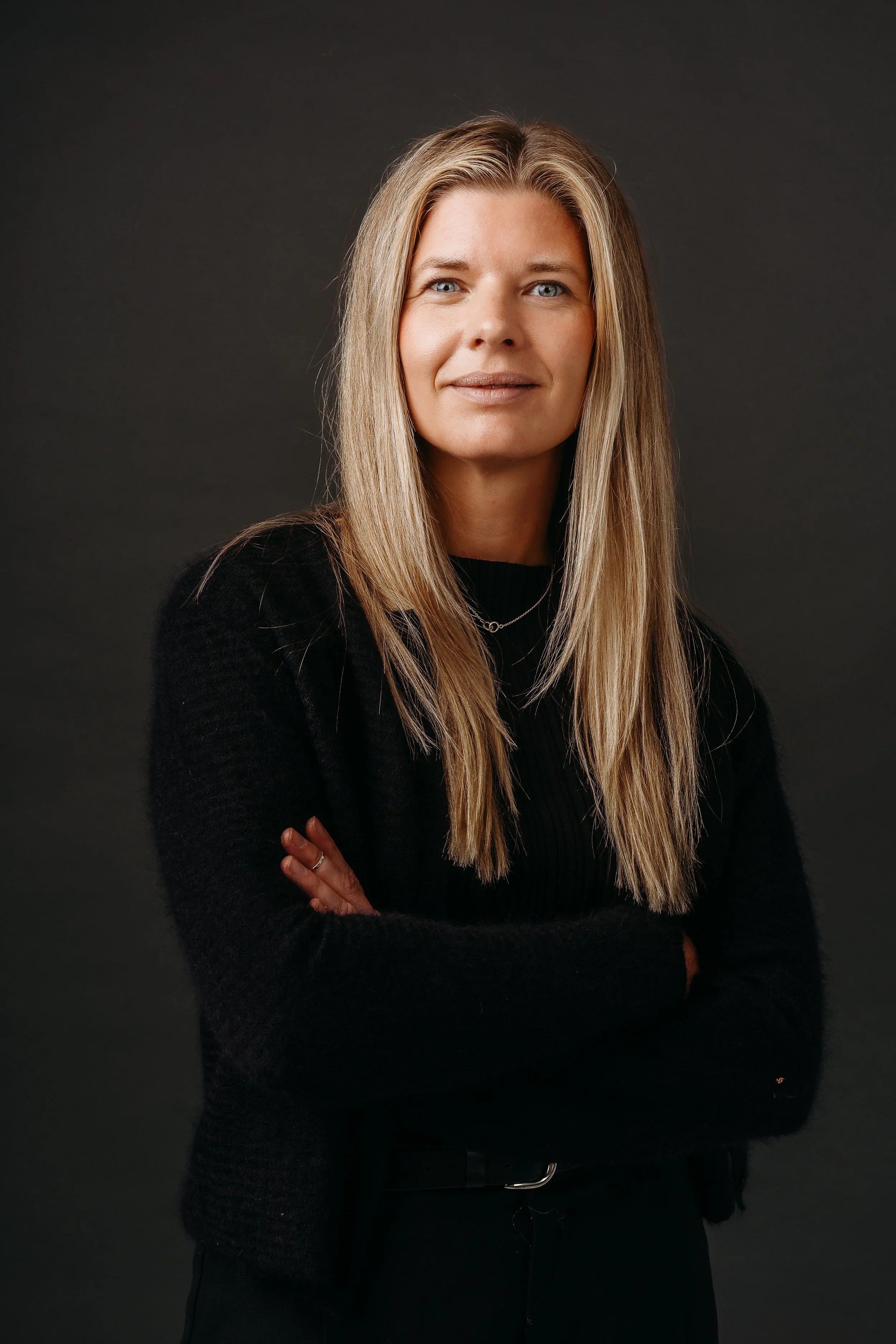 Professional woman with long blond hair, wearing a black top and a necklace, standing with arms crossed against a dark background.