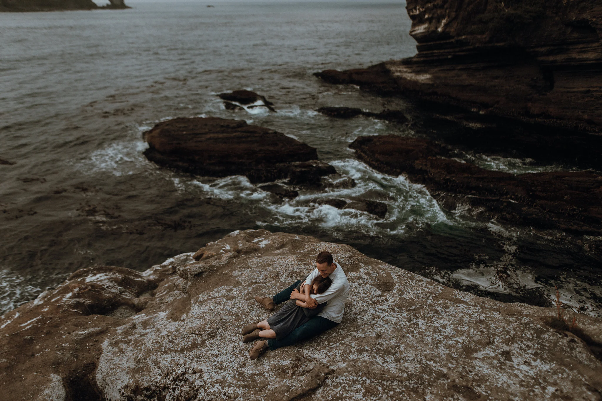 Couple sitting on ocean cliff rock at sunset.