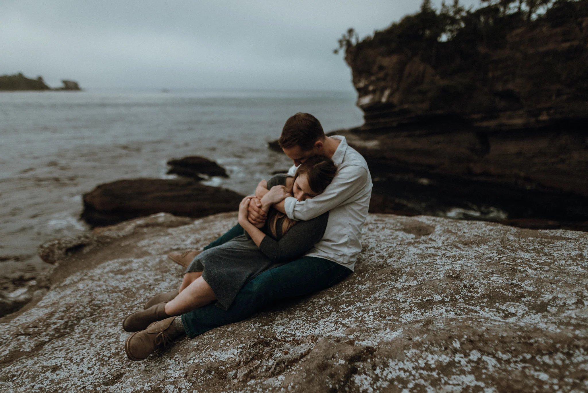 Couple wrapping arms around each other on ocean cliff at sunset.