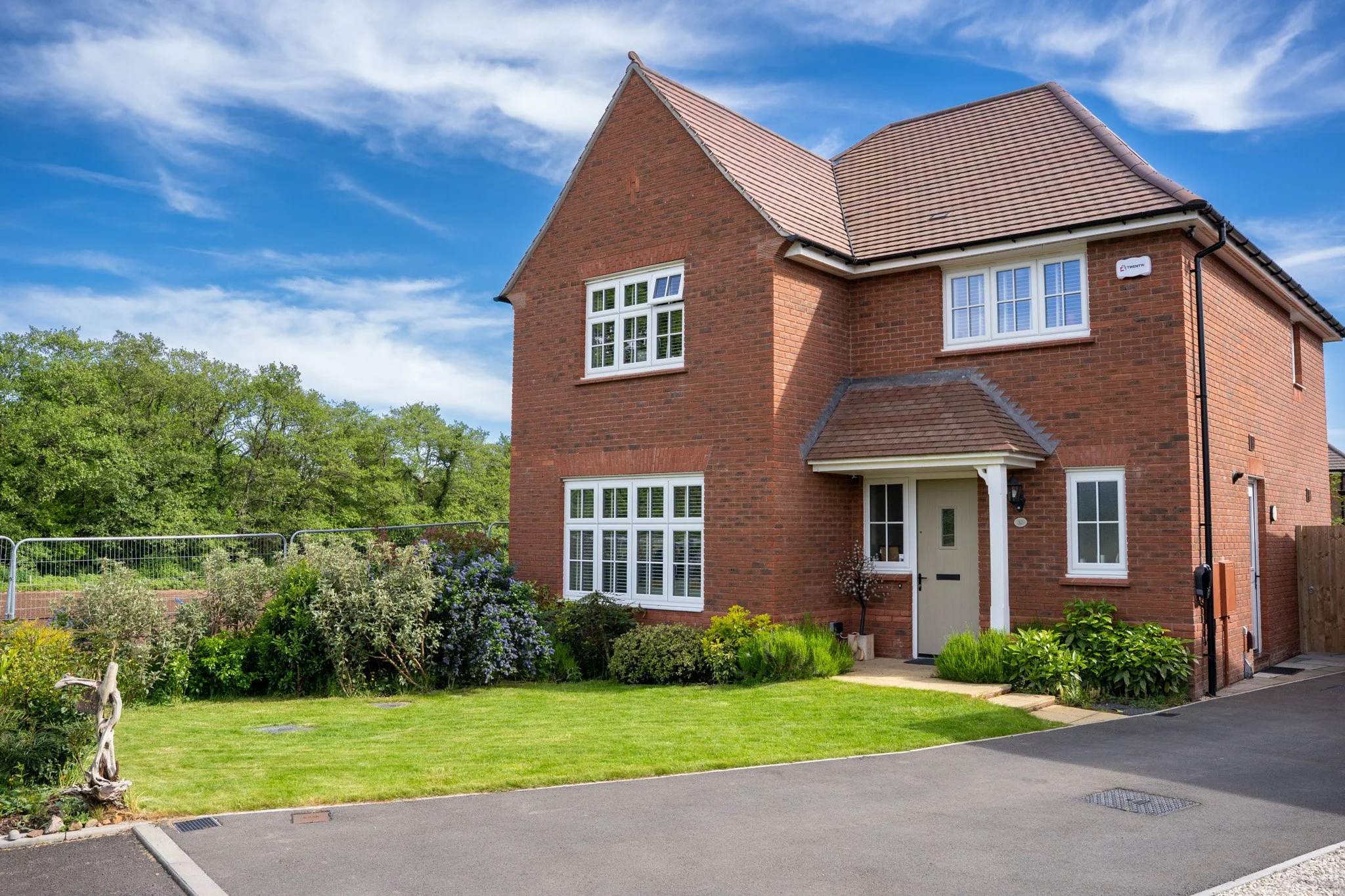 Red brick two-story house with white window frames, surrounded by a garden and green trees under a blue sky with clouds.