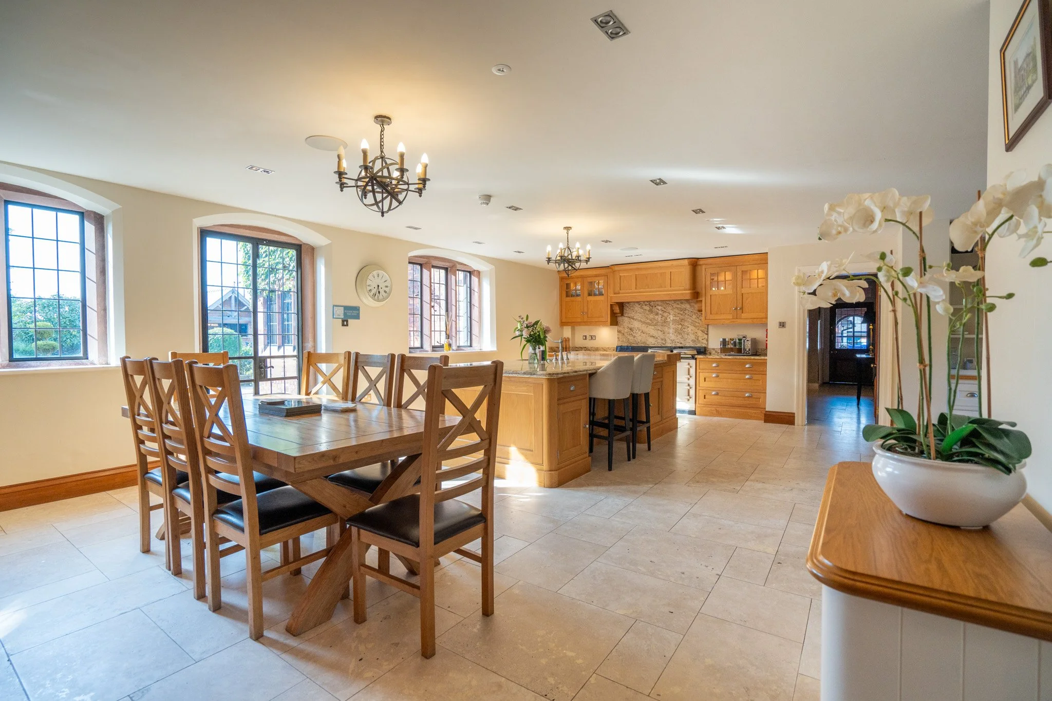 A spacious, well-lit kitchen and dining area with wooden furniture, large windows, pendant chandeliers, and a potted orchid plant.