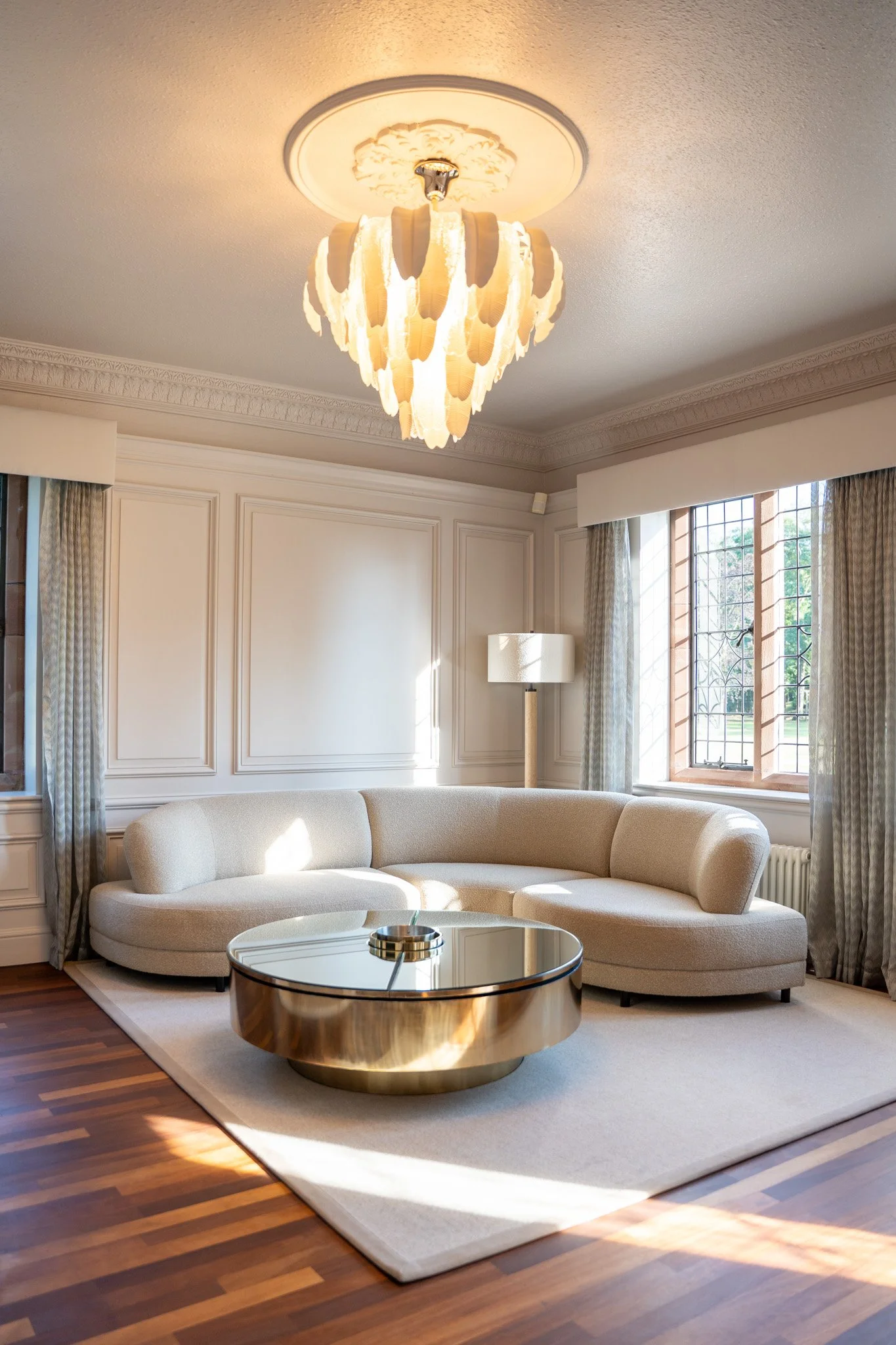 Elegant living room with a cream curved sofa, a round gold coffee table, a white area rug, large window with curtains, and a chandelier on the ceiling.