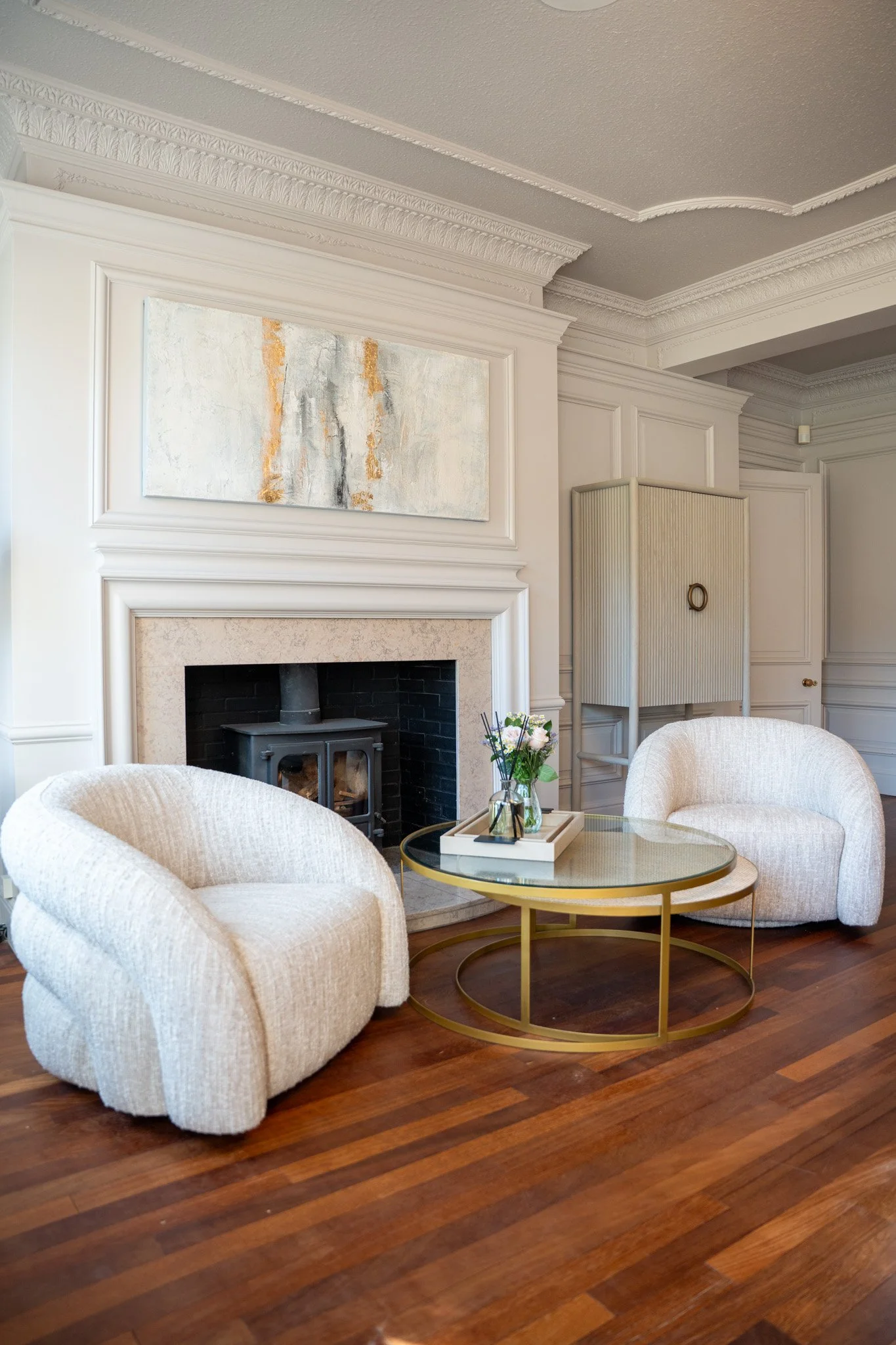 Elegant living room with cream-colored armchairs, a gold circular coffee table, and a decorative abstract painting above a fireplace with black interior. The room has wood flooring and detailed crown molding.