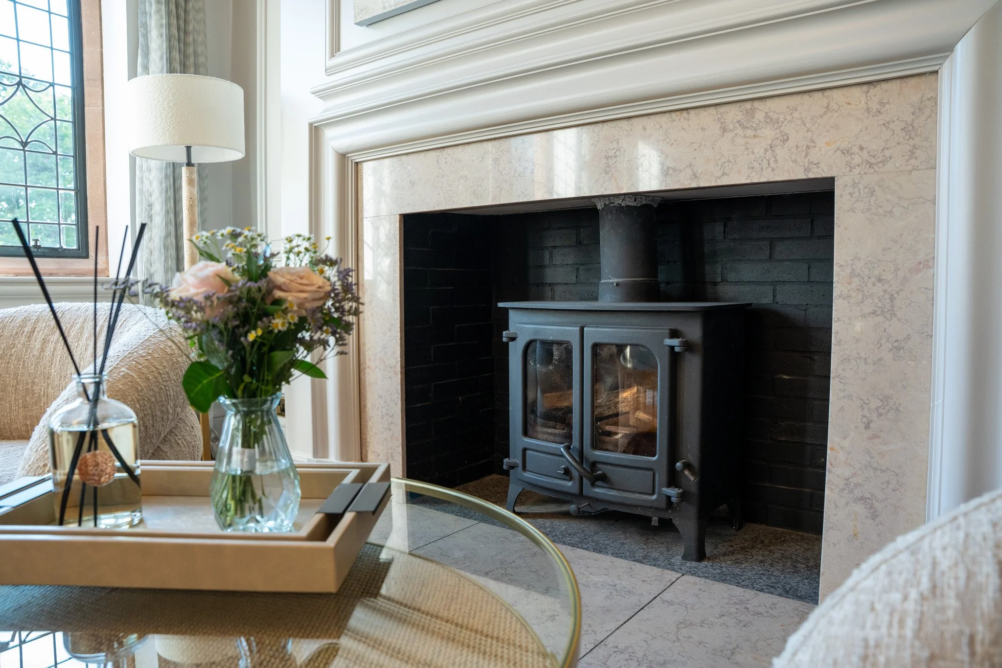 A cozy living room with a black wood stove in front of a marble fireplace, a window with green shutters, a beige sofa, a glass table with a vase of flowers, and a floor lamp in the background.