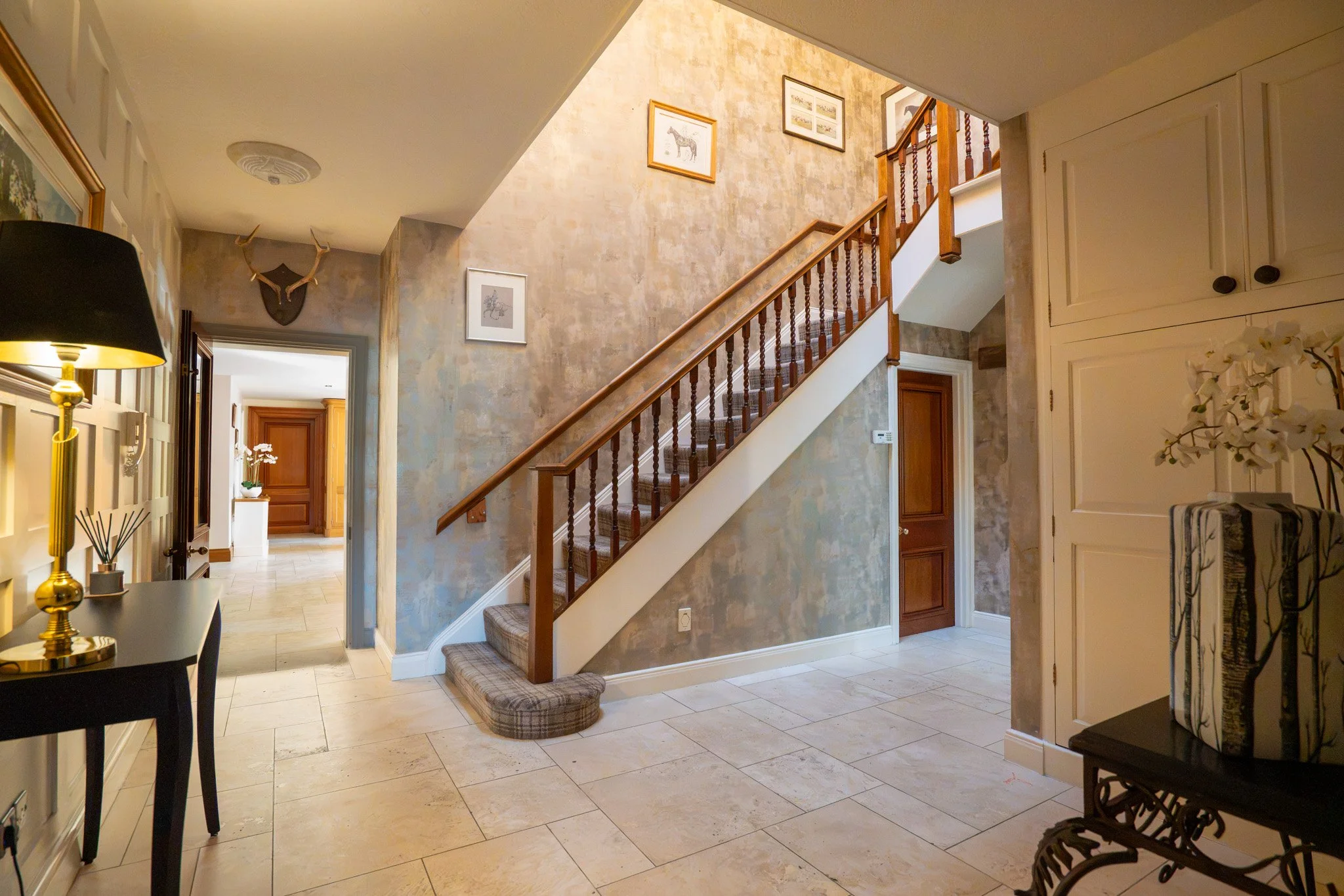 Entryway with tiled floor, staircase with wooden railing, framed pictures on beige textured wall, mounted antlers above doorway, black staircase mat, and a yellow table lamp with a black lampshade.