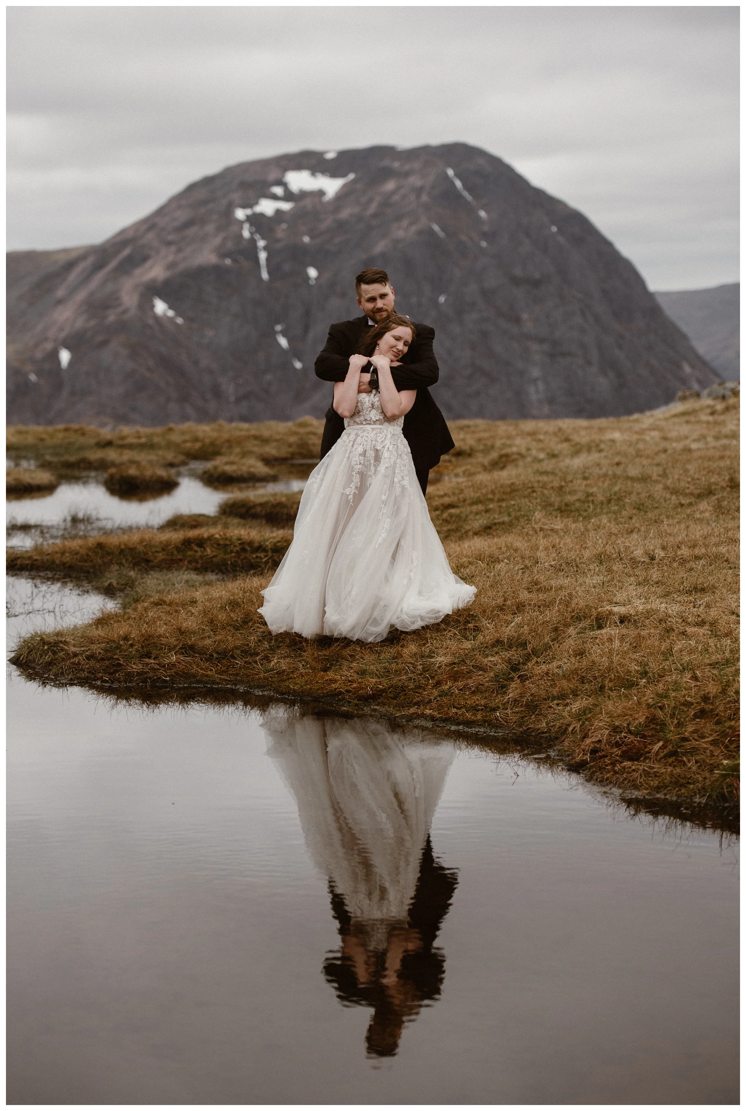 Elissa and Daniel embrace and are reflected in the water a loch in the Scottish Highlands during their destination elopement. Photo by Maddie Mae, Adventure Instead.