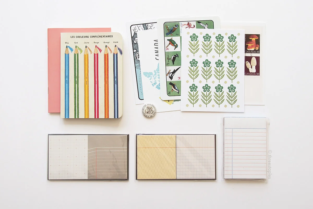 Sticky notes and peach notebook by Paperways , mini letterpressed note pad by Bison Bookbinding & Letterpress , various postcards by The Regional Assembly of Text , and graph grid notebook with color pencils on the cover by Cavallini Papers & Co .