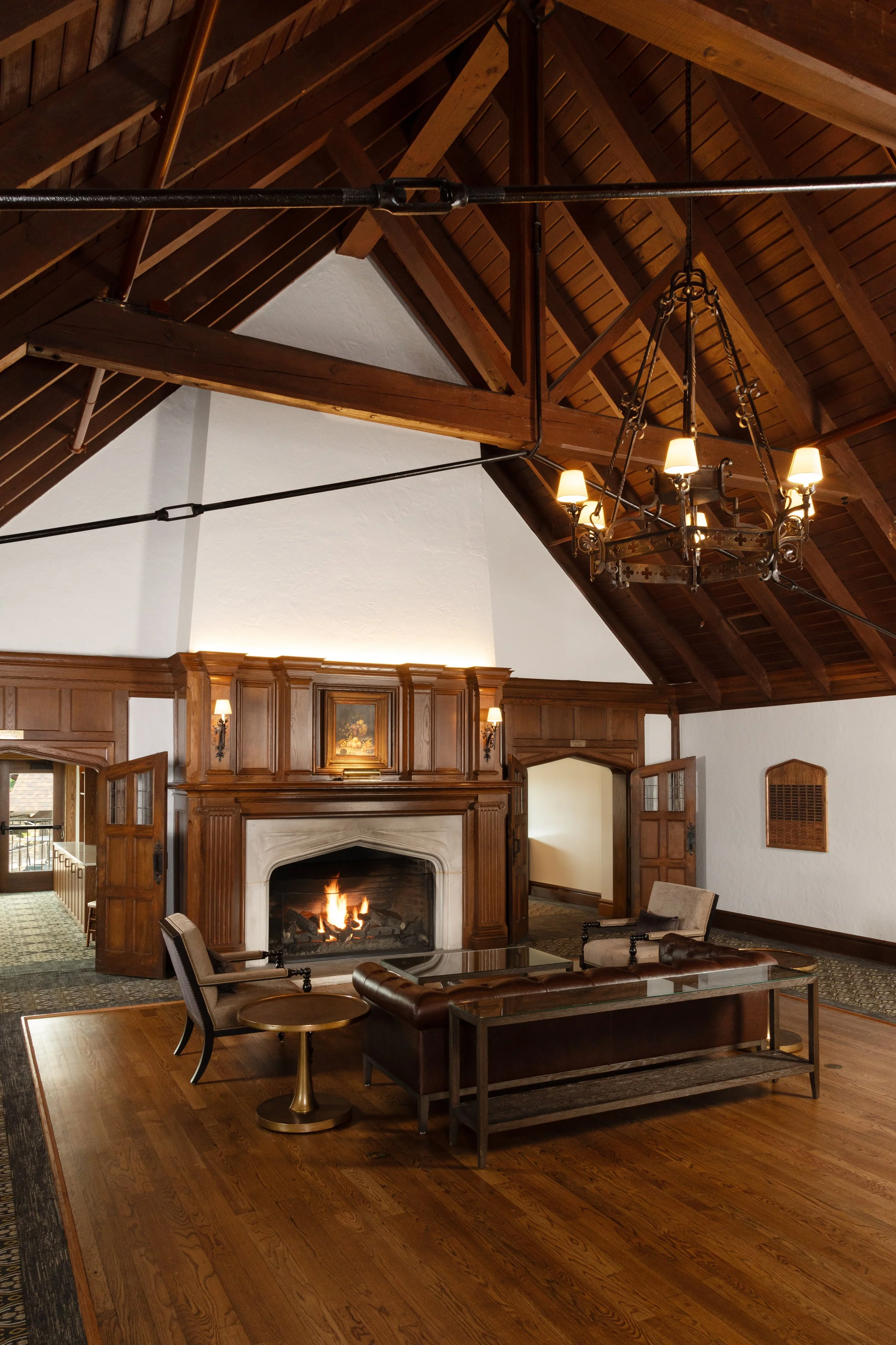 A cozy living room with a wooden vaulted ceiling, chandelier, white walls, a fireplace with a burning fire, and vintage furniture including a sofa, chairs, and side tables.