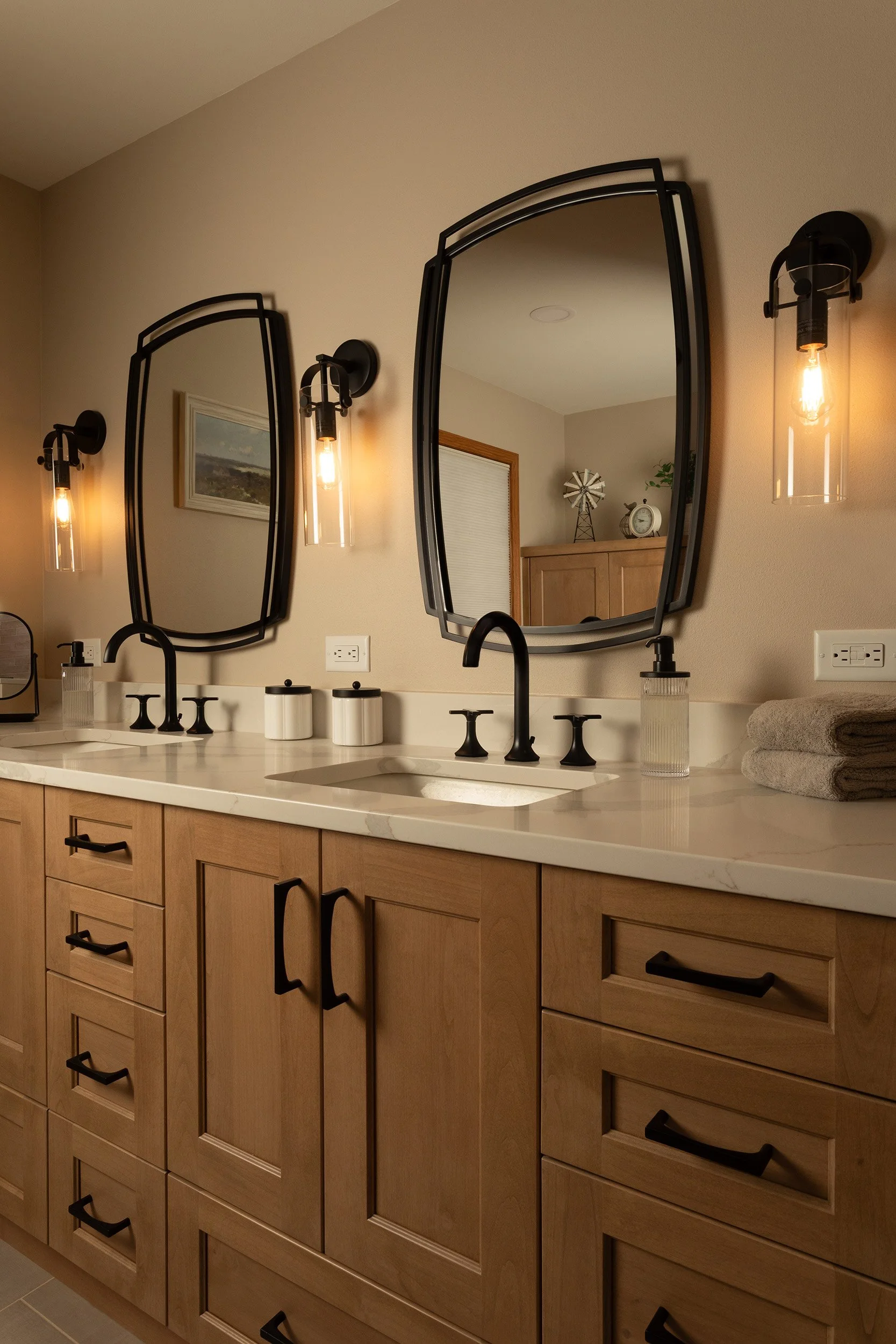 A bathroom vanity with light wood cabinets, black handles, two black sinks, and a marble countertop. Above, three large mirrors are flanked by two black wall-mounted light fixtures with Edison bulbs. Towels are folded on the right side of the counter