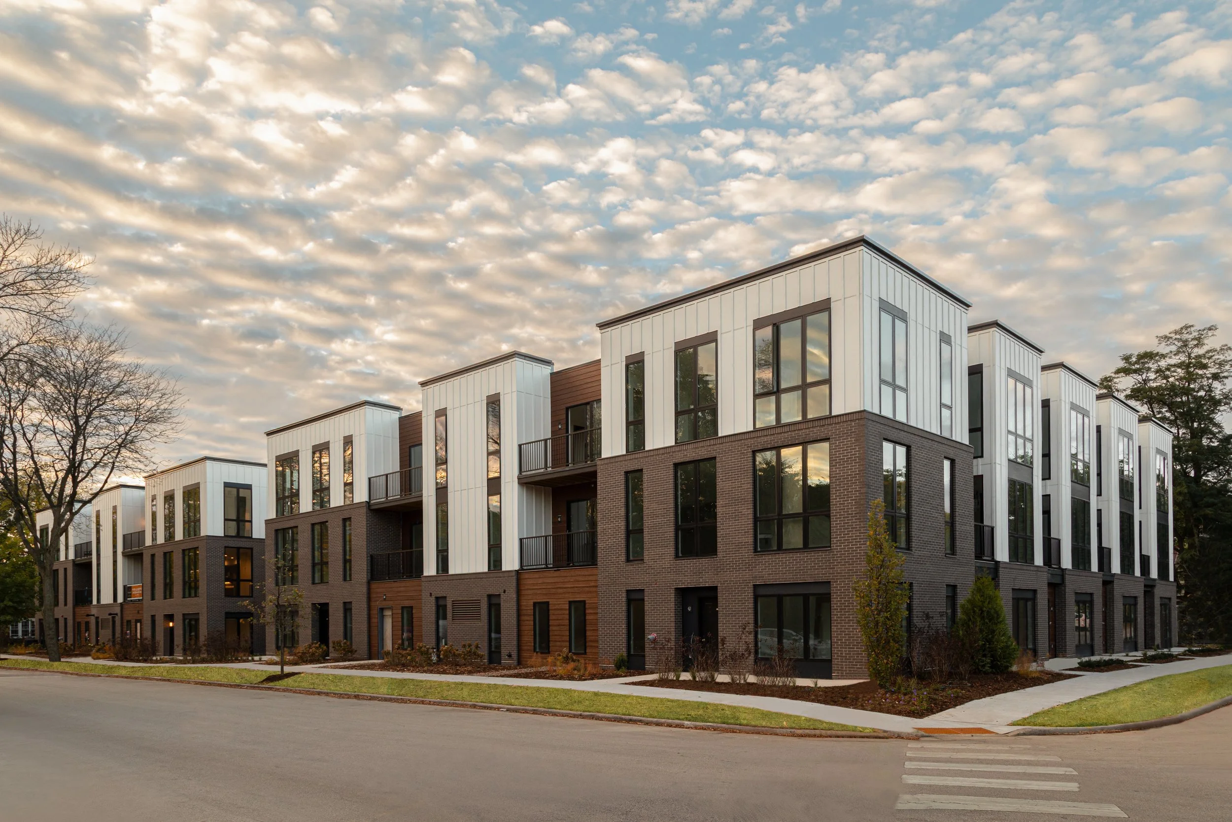 Modern multi-story apartment building with large windows and balconies, set against a partly cloudy sky, with trees and a sidewalk in the foreground.