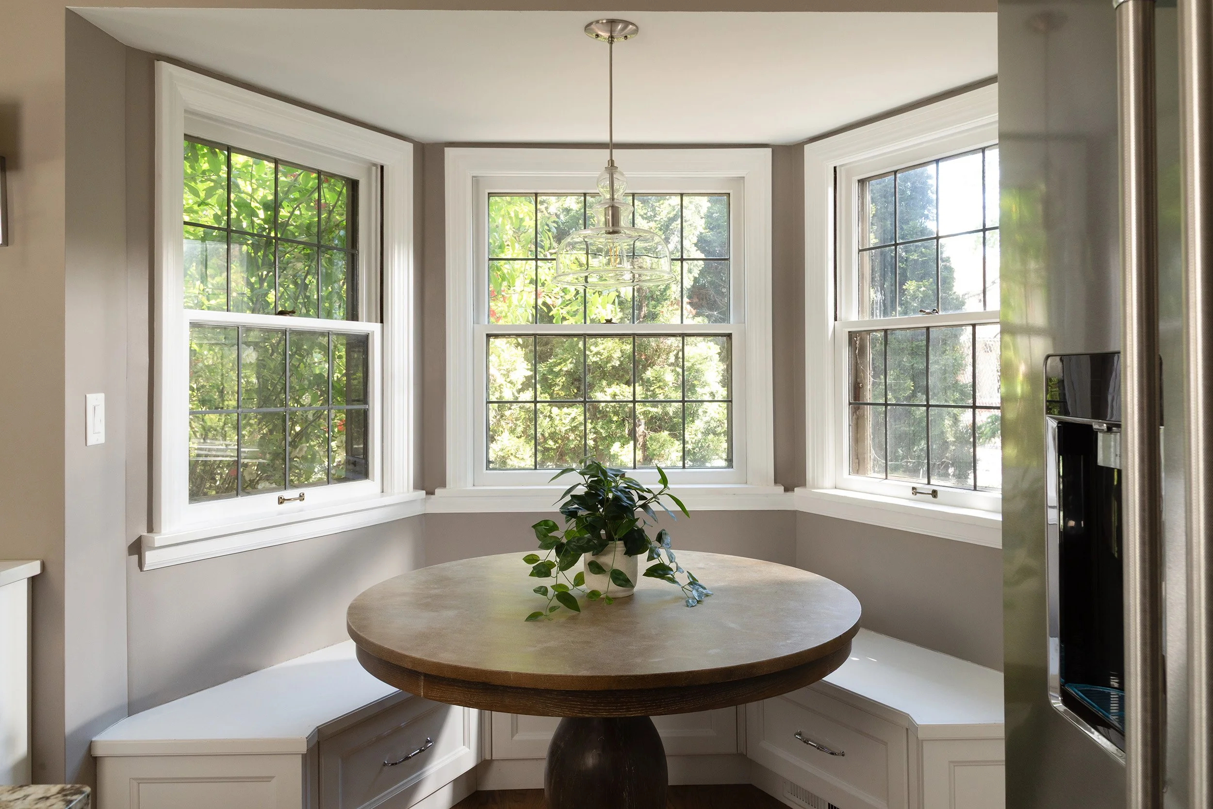 A kitchen nook with three large windows, a round wooden table with a potted plant, and a hanging light fixture.