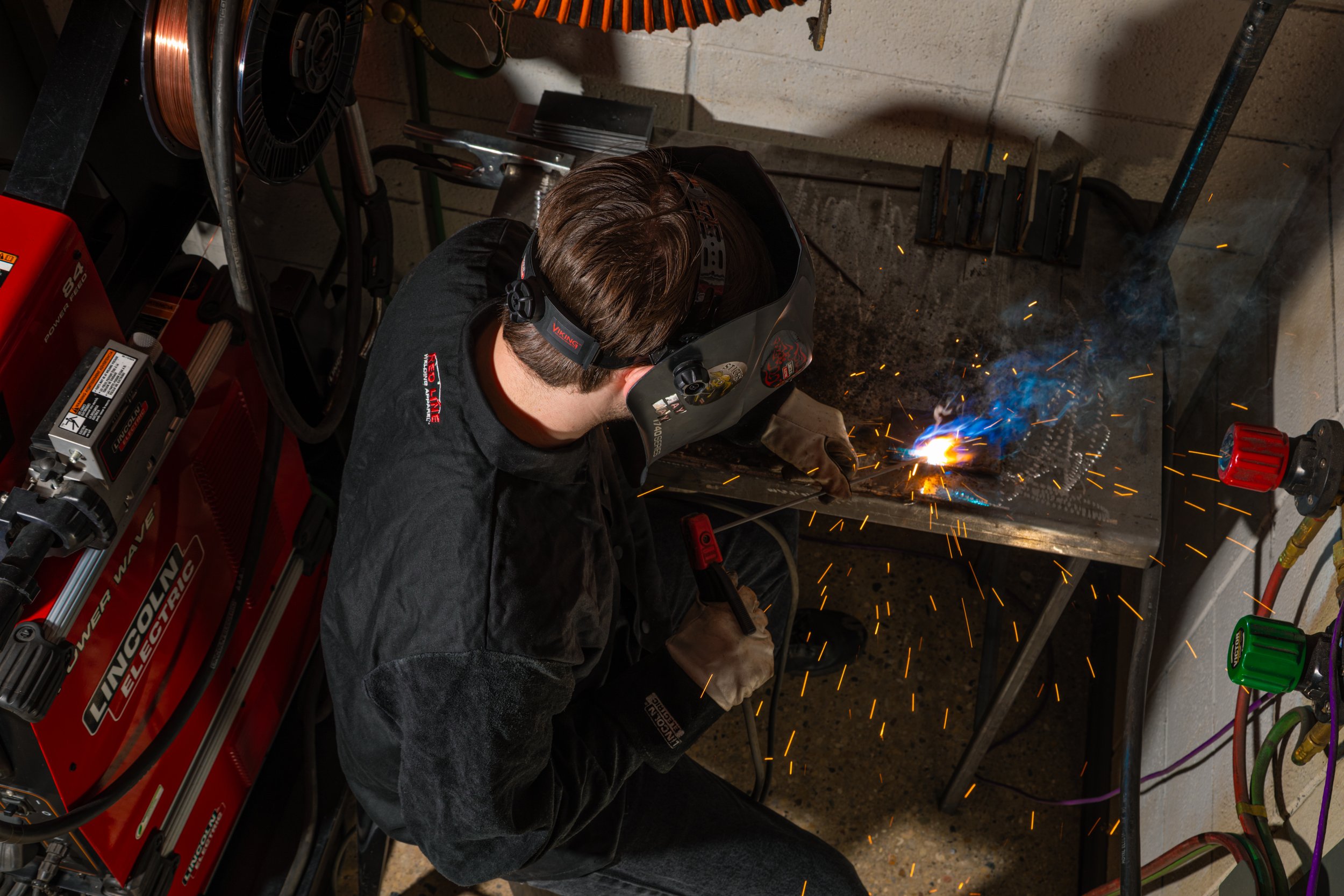 A person welding metal in a workshop, wearing a welding helmet and protective gloves, with sparks flying from the welding process.