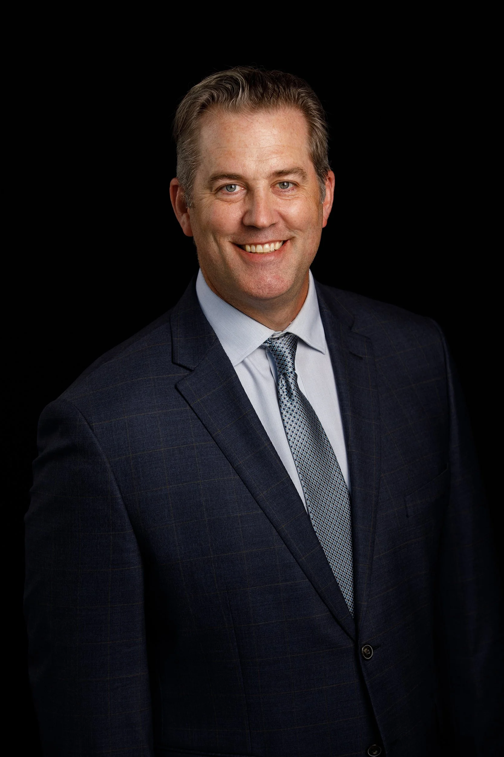  Photography by Tom Krueger, photographer,  Milwaukee. A professional headshot of a smiling middle-aged man with light-colored eyes and brown hair, wearing a dark checkered suit with a light shirt and patterned tie, against a black background.