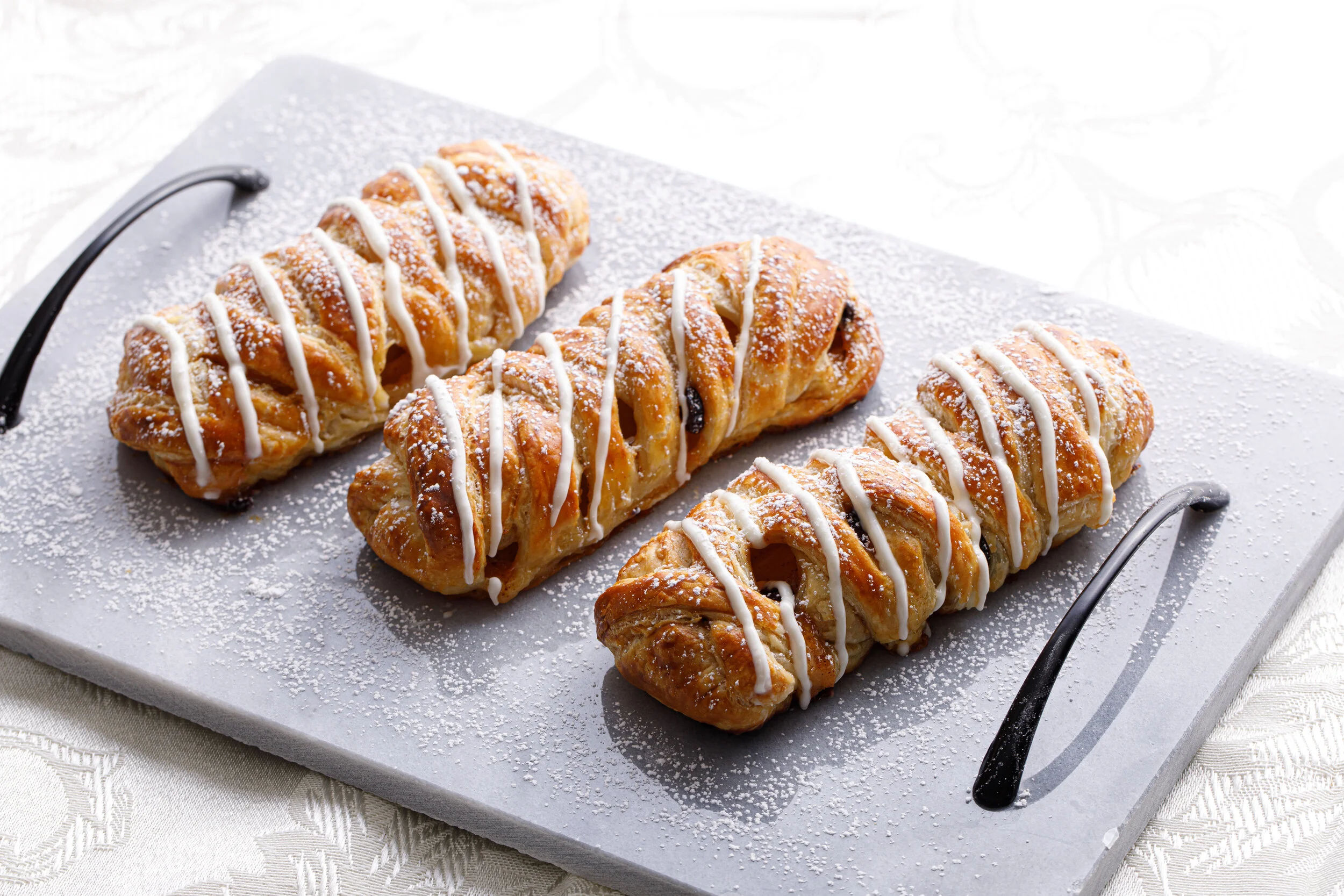 Three pastry twists topped with powdered sugar and drizzled with white icing, arranged on a serving tray.