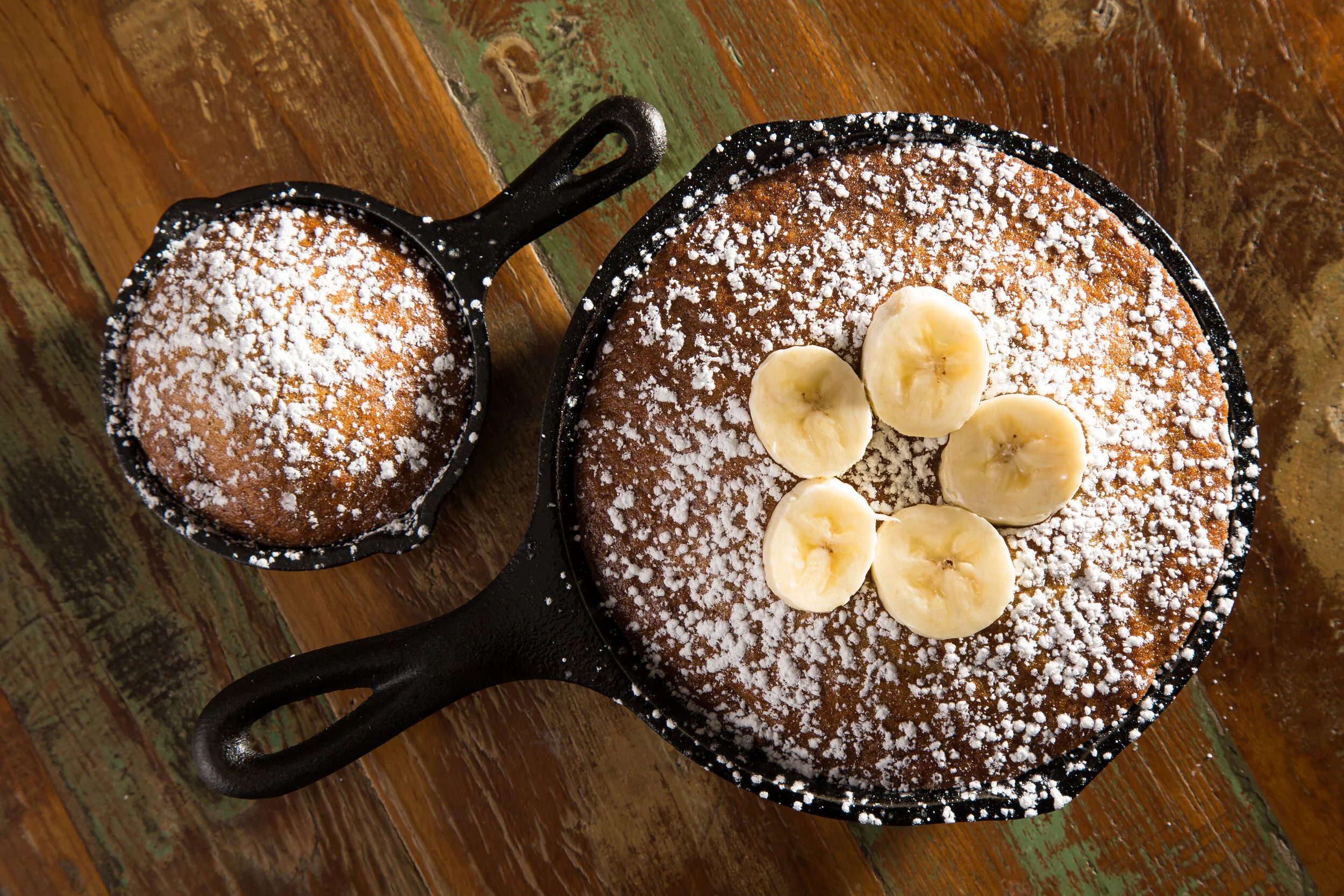 A round banana cake topped with powdered sugar and banana slices in a cast iron skillet. A smaller cast iron skillet beside it contains a round cake dusted with powdered sugar.