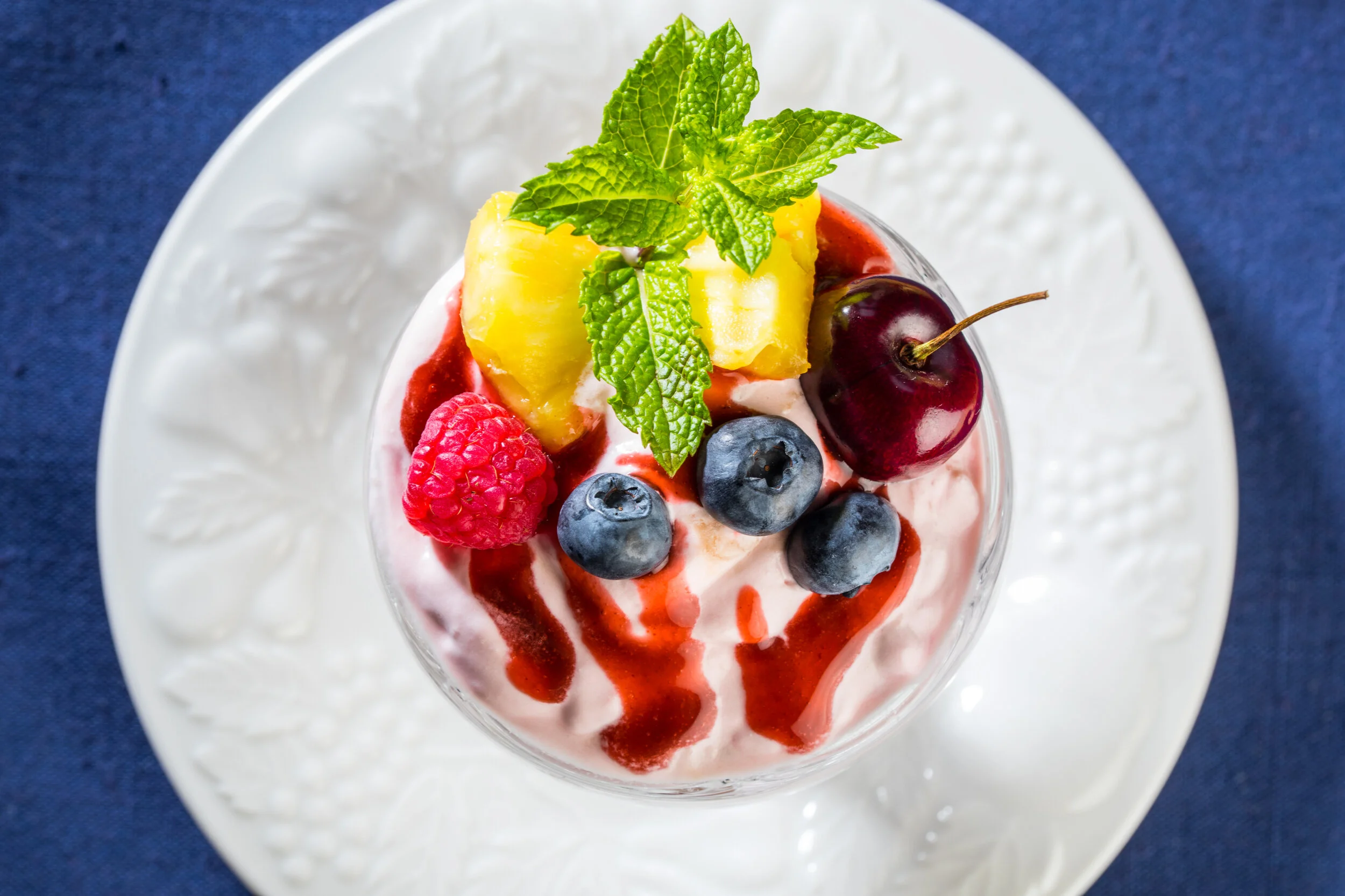 Dessert in a glass topped with pineapple chunks, cherry, raspberries, blueberries, and mint leaves, with a swirl of fruit sauce.