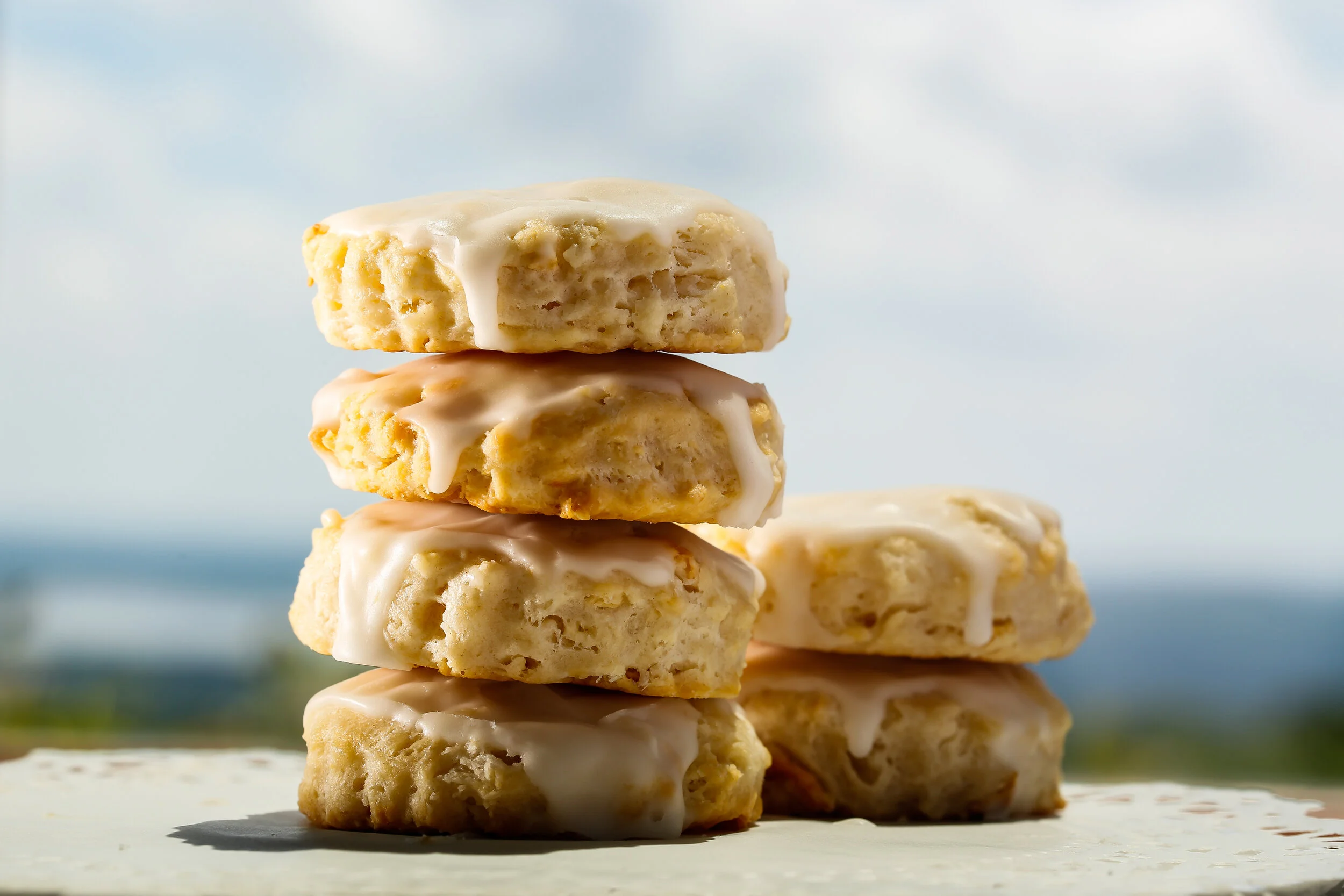 Stacked cookies with white icing on a white surface, outdoors with a cloudy sky and blurred landscape in the background.