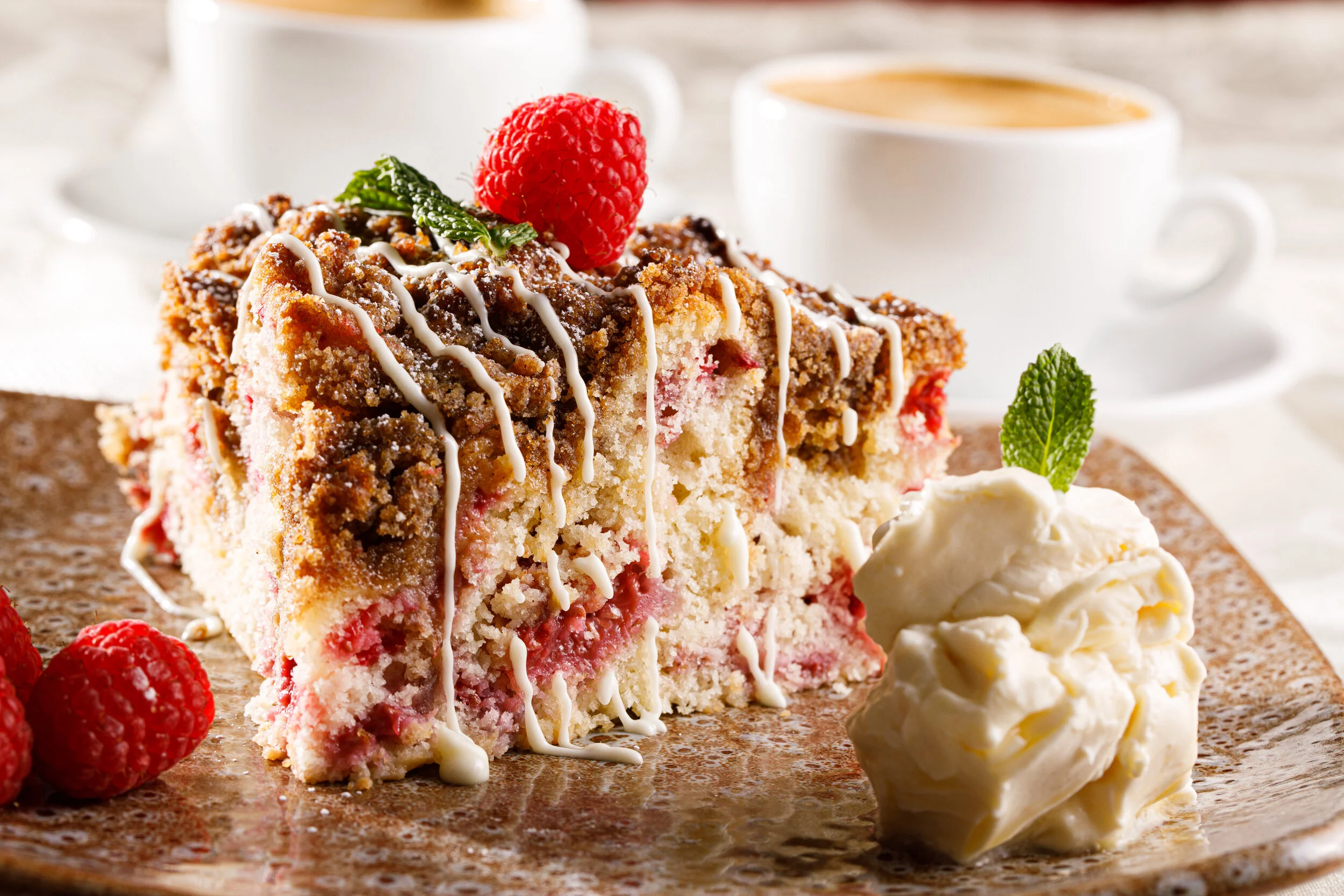 Slice of raspberry crumb cake with white icing drizzle, fresh raspberries, and a dollop of whipped cream with mint, served with coffee in cups in the background.